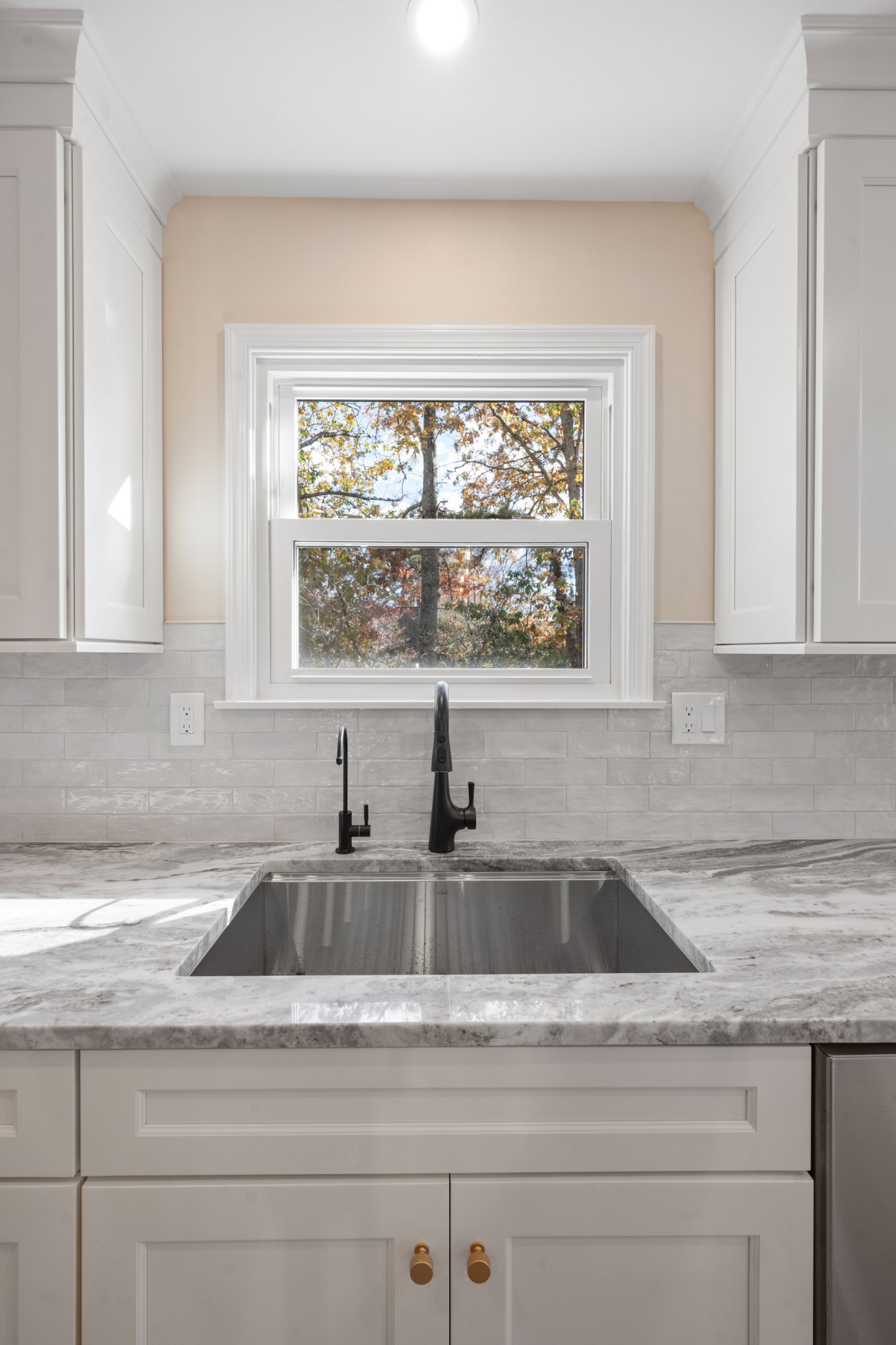 A modern kitchen features a marble countertop with a large double sink, black faucet, white cabinets, and a window above the sink letting in natural light and showing trees outside.