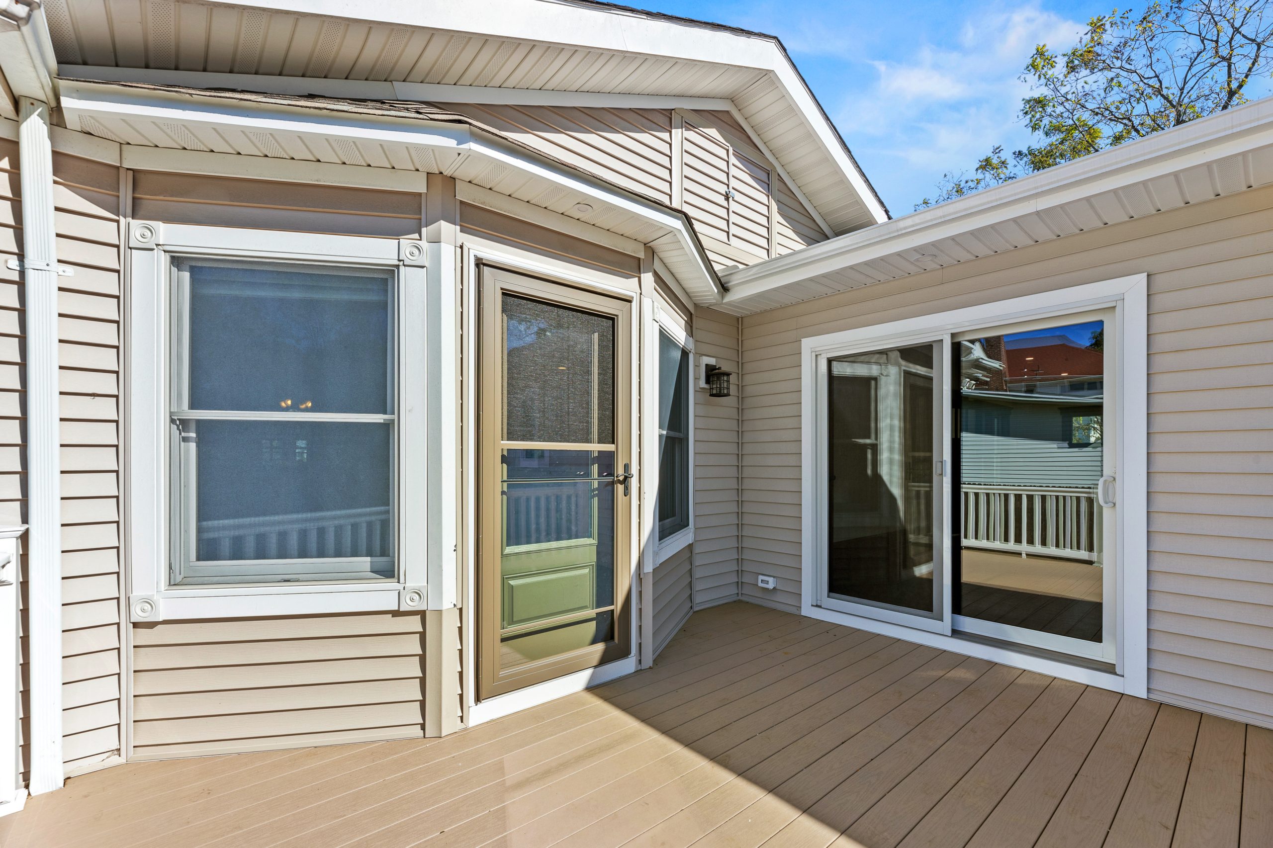 A beige house with light siding, featuring a glass storm door, two large windows, and a sliding glass door opening onto a spacious, light brown wooden deck under a clear blue sky.
