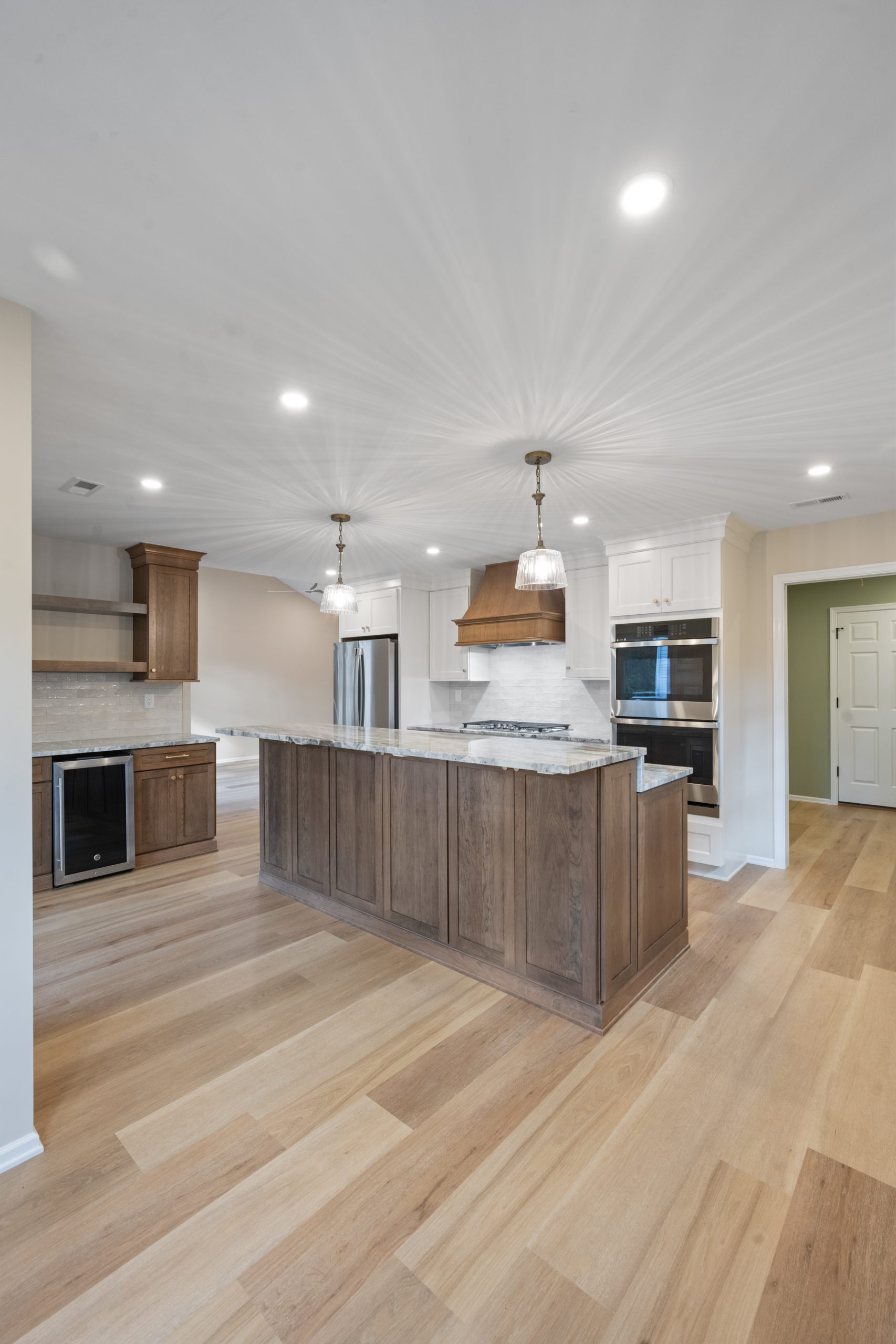 Modern kitchen with light wood floors, a large island with dark wood cabinetry, pendant lights, marble countertops, stainless steel appliances, double ovens, open shelving, and a wine fridge. Neutral walls and bright overhead lighting.