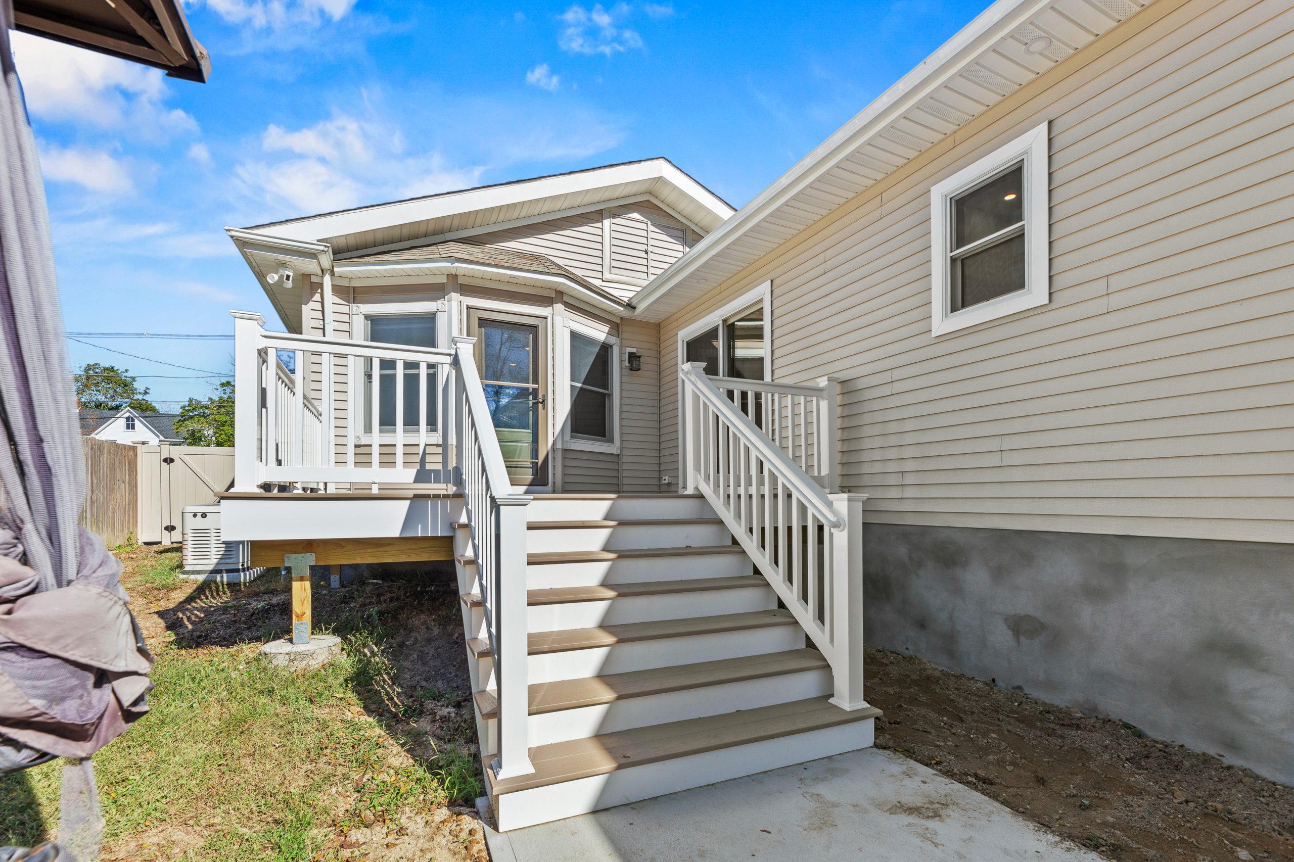 A small beige house with white trim features a raised wooden deck and stairs leading to a back door. The yard is partially bare, and the sky is blue with a few clouds.