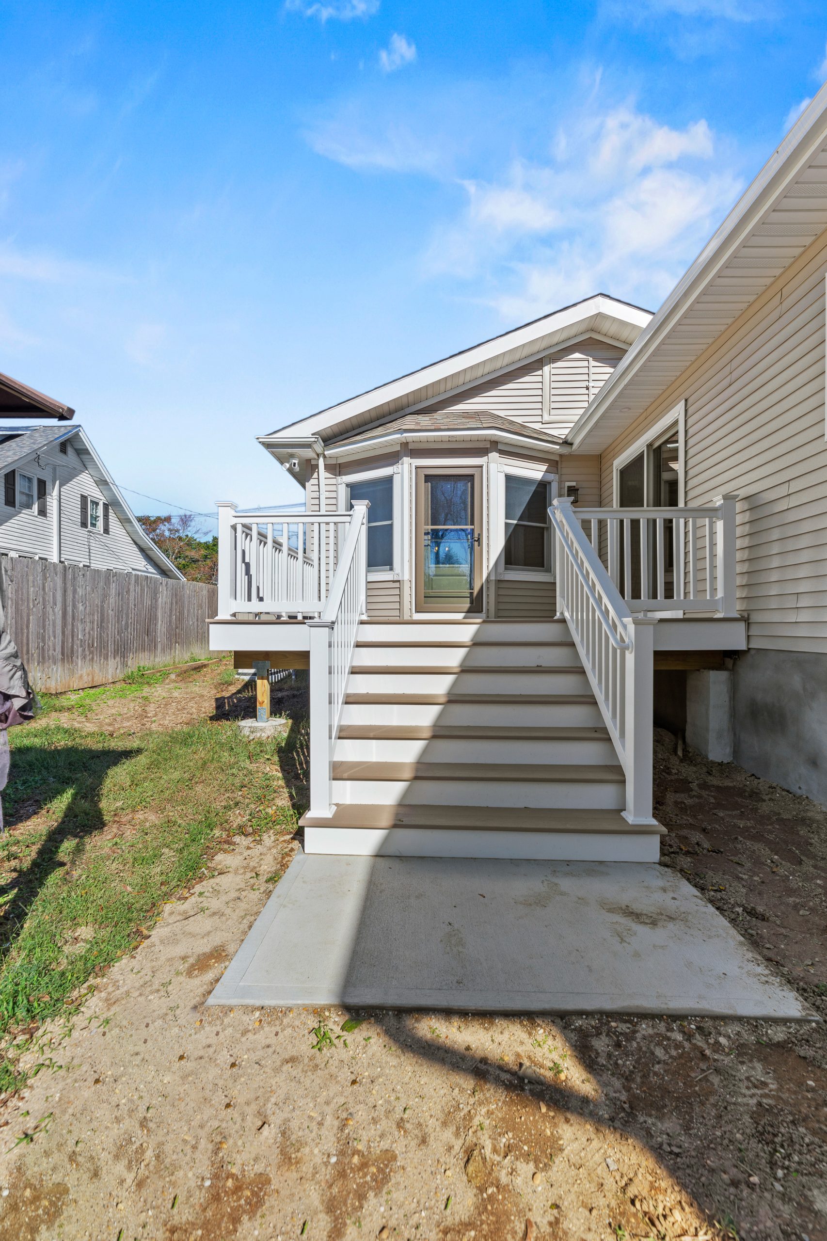 A back view of a house showing a set of white steps with railings leading up to a small porch and a glass door. The yard has patches of grass and dirt, and the sky is blue with some clouds.