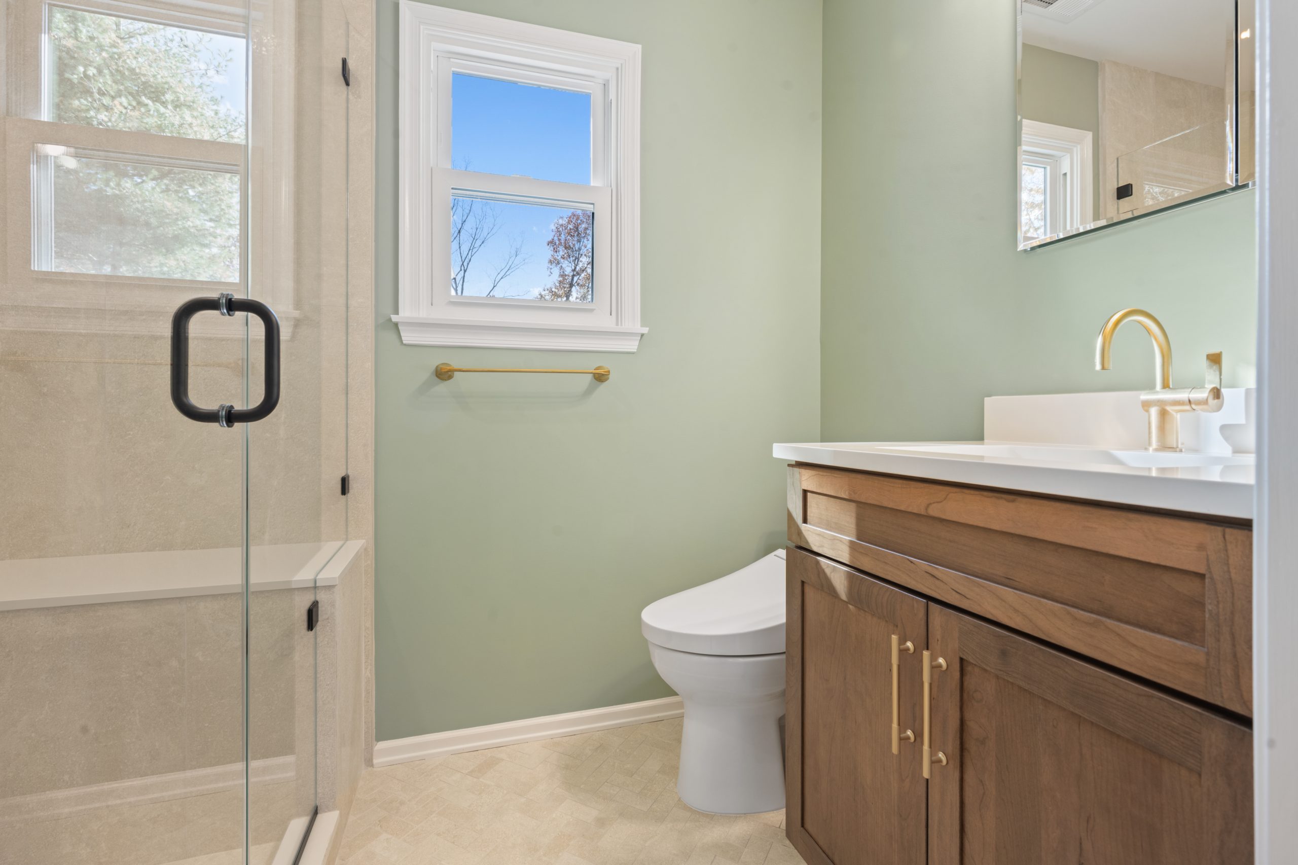 A modern bathroom with light green walls, a glass shower, gold fixtures, a white toilet, a wooden vanity with a white countertop, and two small windows letting in natural light.