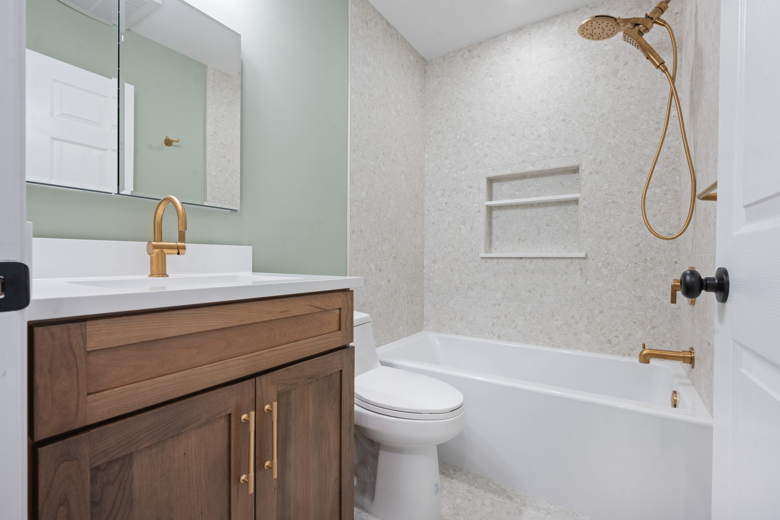 Modern bathroom with a wooden vanity, white countertop, gold fixtures, a toilet, and a bathtub with a built-in shelf and gold showerhead; light-colored walls and floor create a clean, bright look.