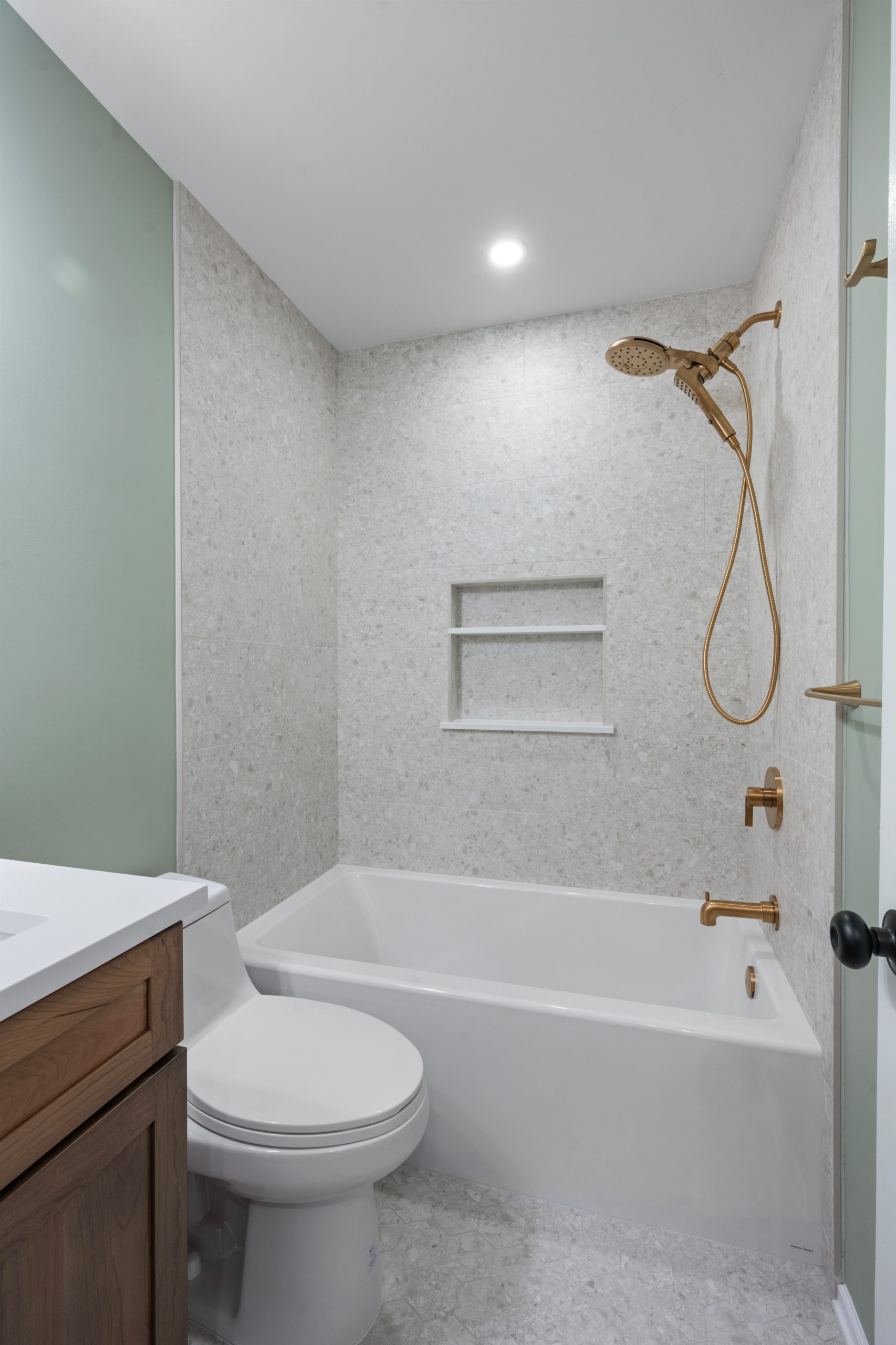 Modern bathroom with a white bathtub, built-in wall niche, gold showerhead and faucet, white toilet, wood vanity, and light gray textured walls and floor. Ceiling has a recessed light.