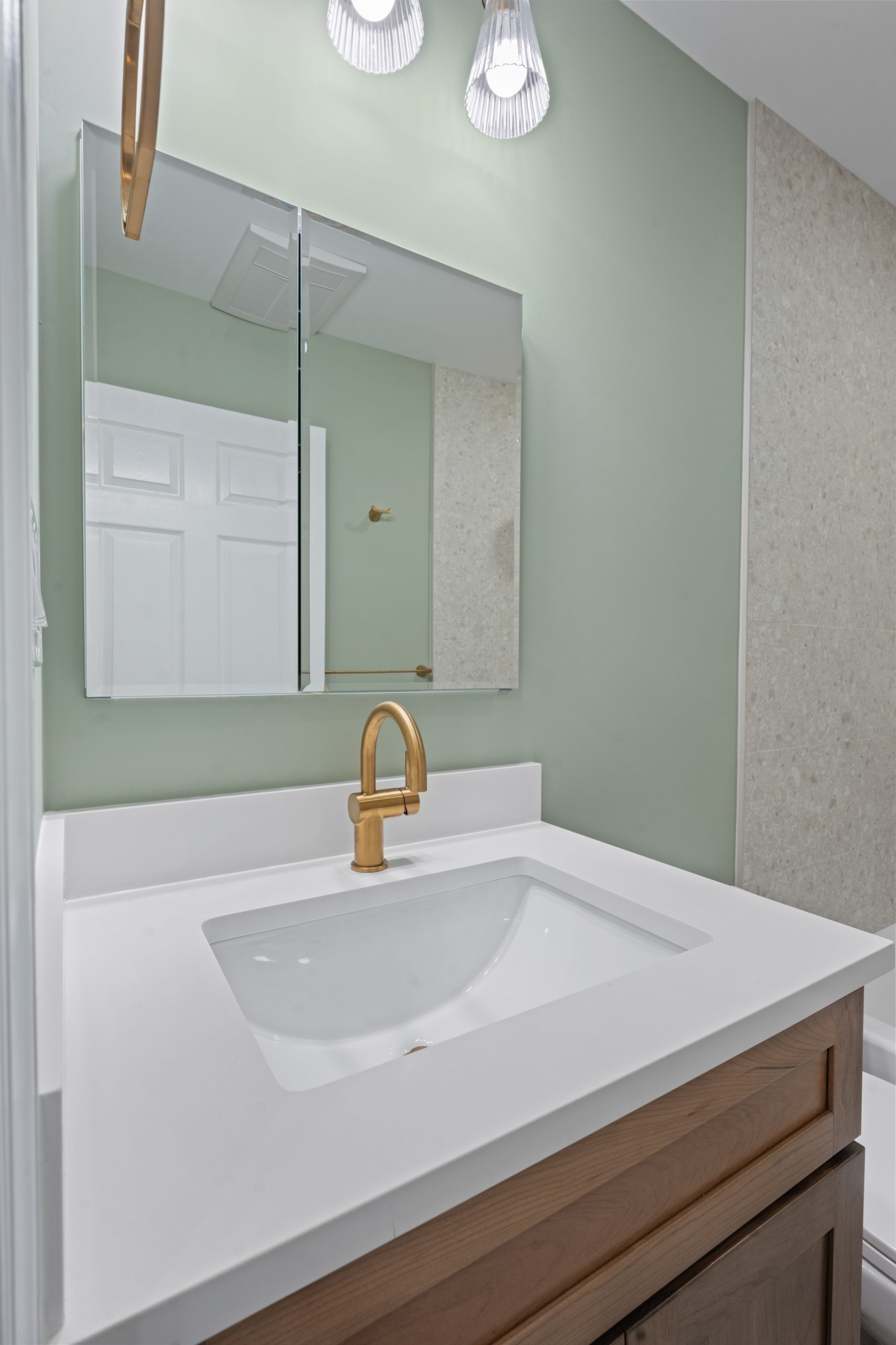 A bathroom vanity with a white countertop, rectangular sink, gold faucet, wooden cabinet, and a mirrored medicine cabinet above. The wall is painted light green and two light fixtures are mounted above the mirror.