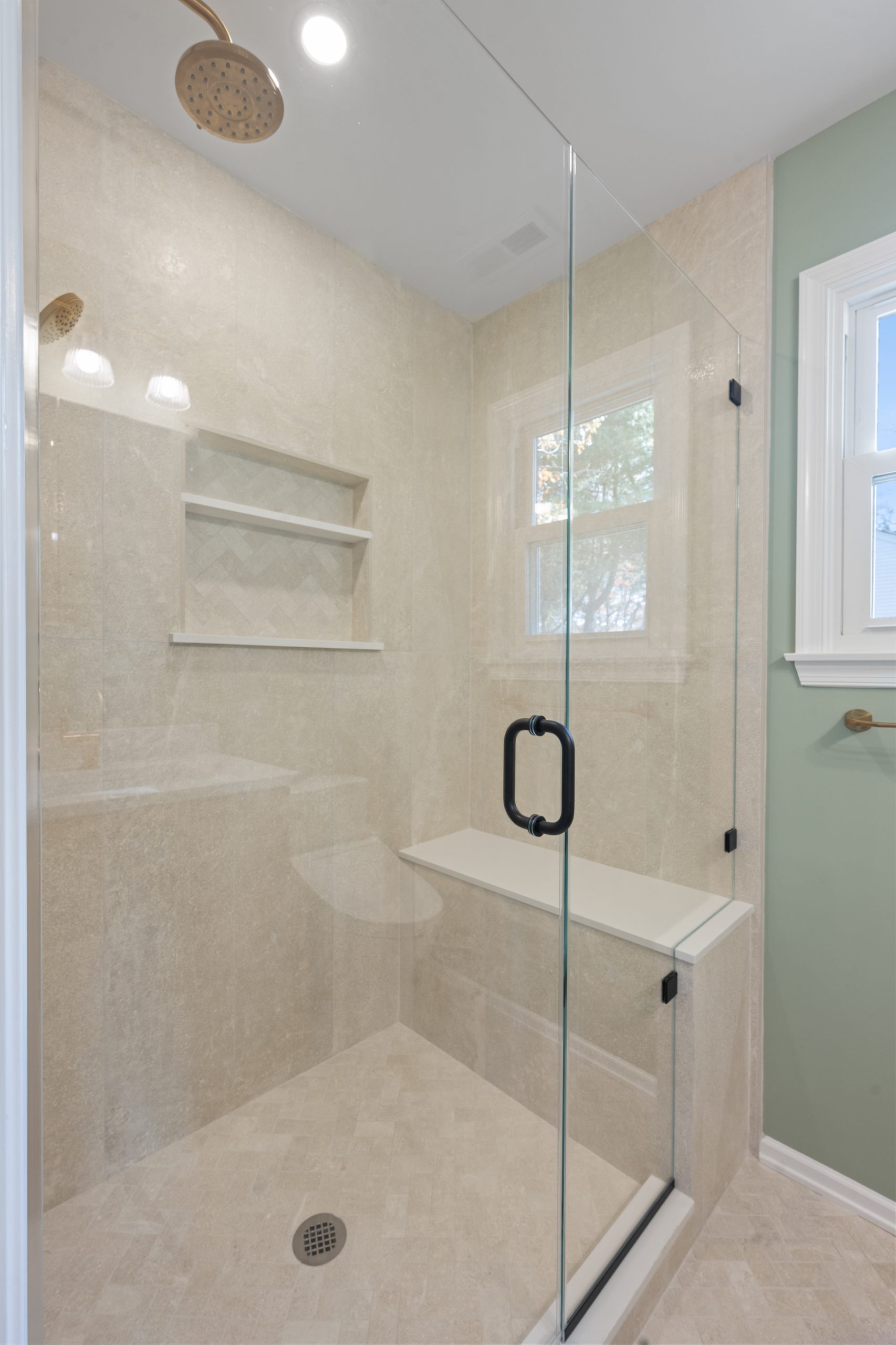 A modern glass-enclosed shower with a built-in bench, two wall shelves, beige tile walls and floor, a gold showerhead, and a window letting in natural light.