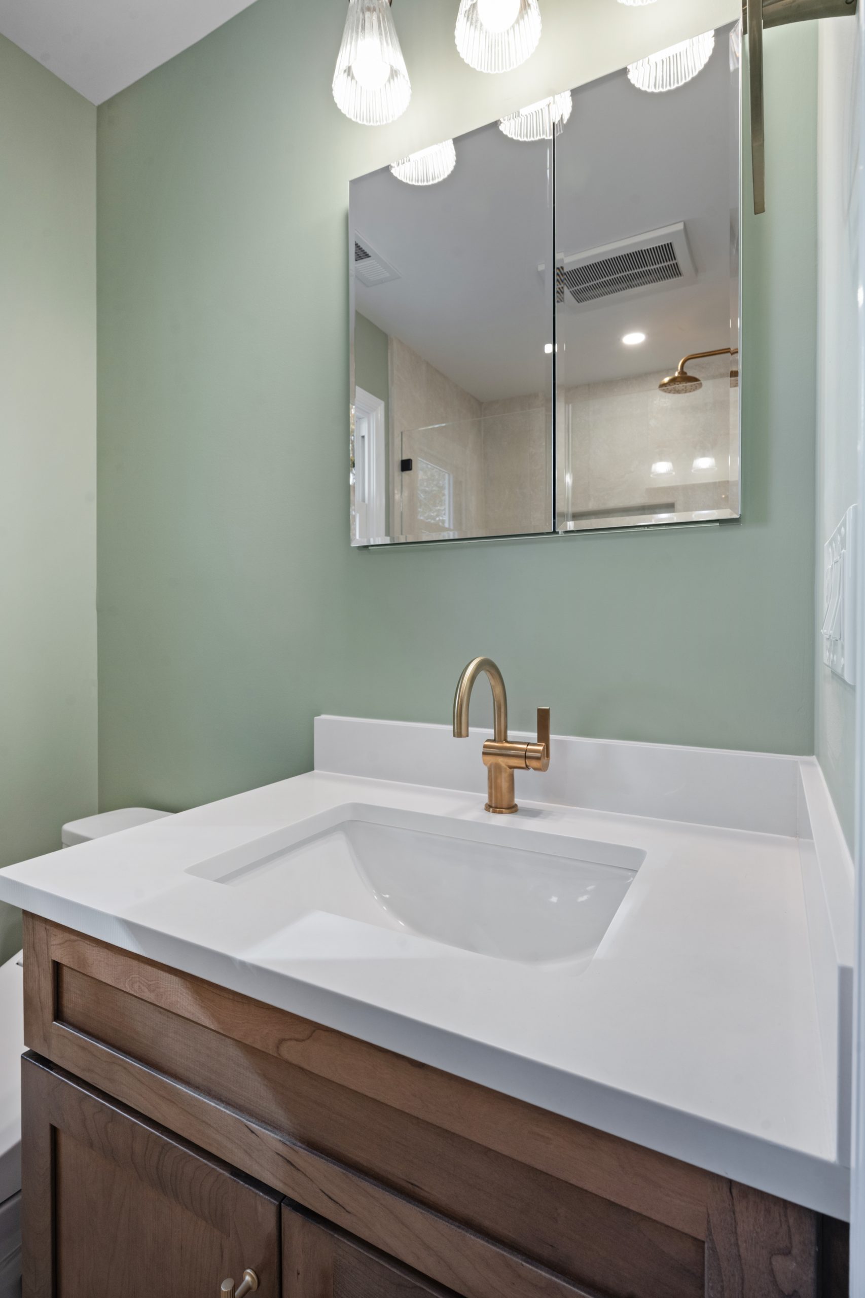 A modern bathroom vanity with a white countertop, gold faucet, wooden cabinet, mirrored medicine cabinet, and sage green wall, with light fixtures above the mirror.