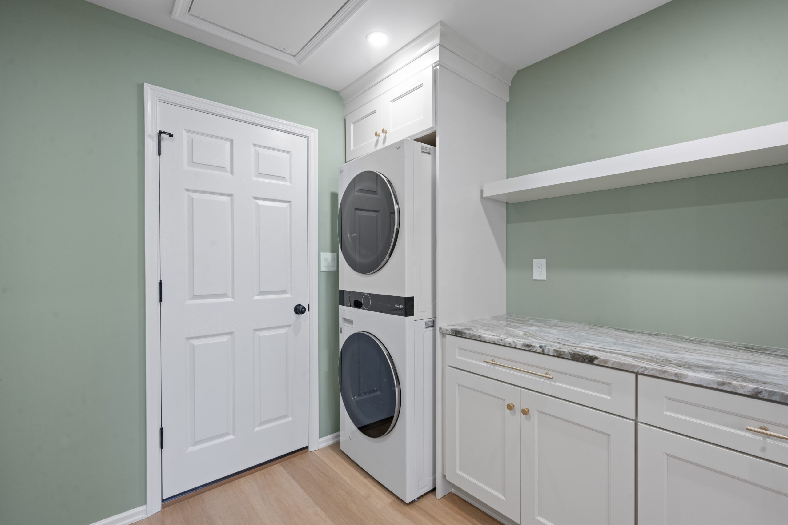 A modern laundry room with green walls, a white door, stacked washer and dryer, white cabinets with gold handles, and a gray marble countertop.