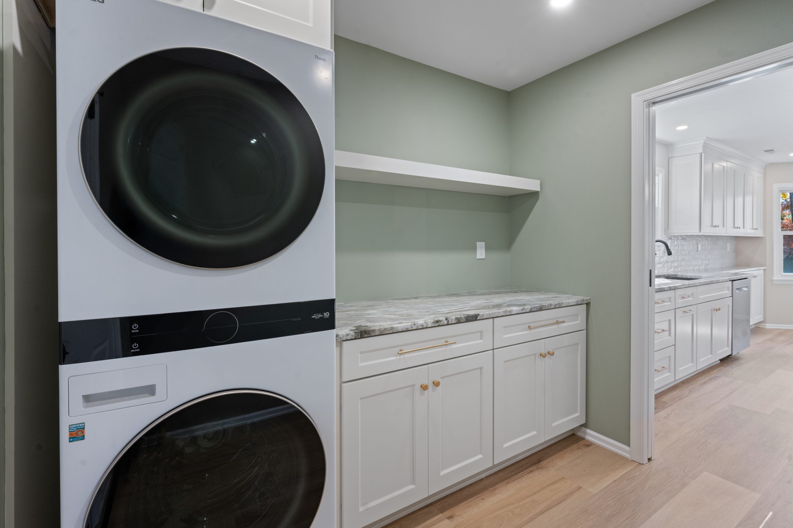 A modern laundry room with a stacked washer and dryer on the left, white cabinets with gold handles, a marble countertop, green walls, and an open doorway to a bright kitchen with white cabinetry.