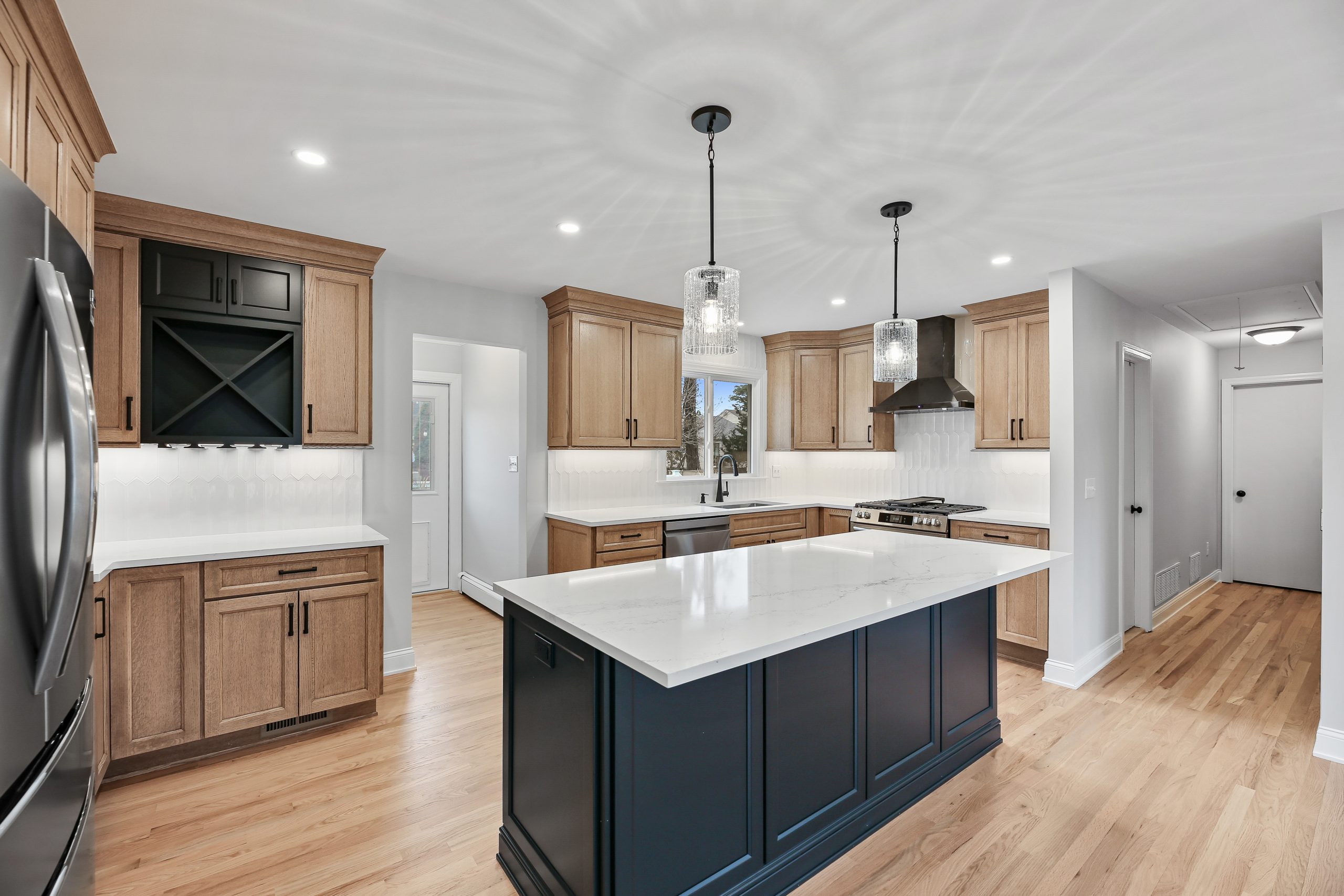 Modern kitchen with wood cabinets, a large white marble island with dark blue base, stainless steel appliances, two pendant lights, light wood flooring, and white tile backsplash. Ample lighting brightens the space.
