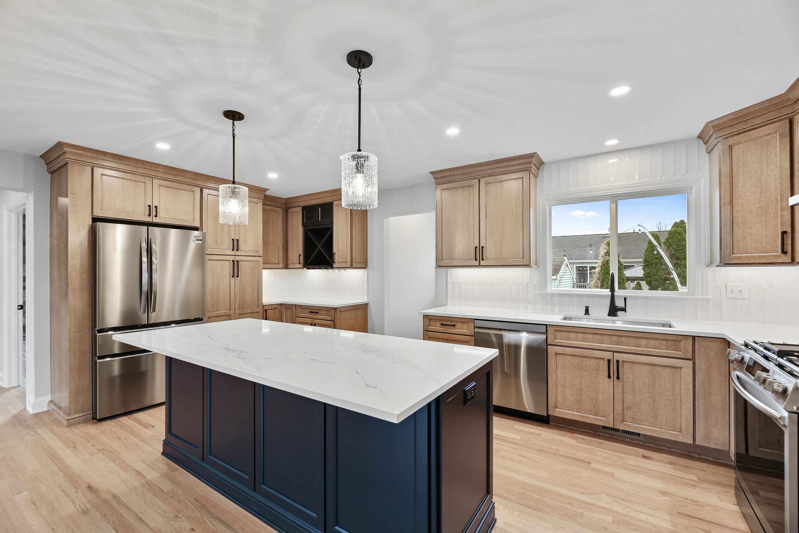 Modern kitchen with light wood cabinets, stainless steel appliances, a navy blue island with a white marble countertop, pendant lights, wood floors, and a large window over the sink.