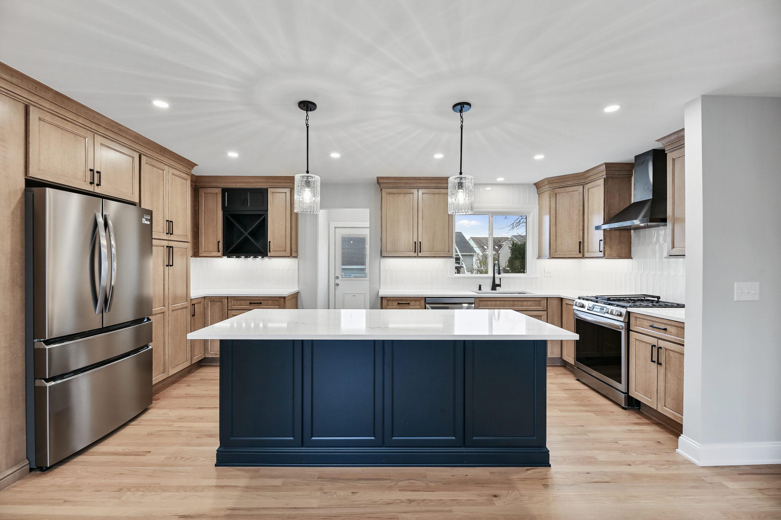 Modern kitchen with wood cabinets, stainless steel appliances, a large navy blue island with a white countertop, two pendant lights above the island, and hardwood floors. White tile backsplash and ample natural light.