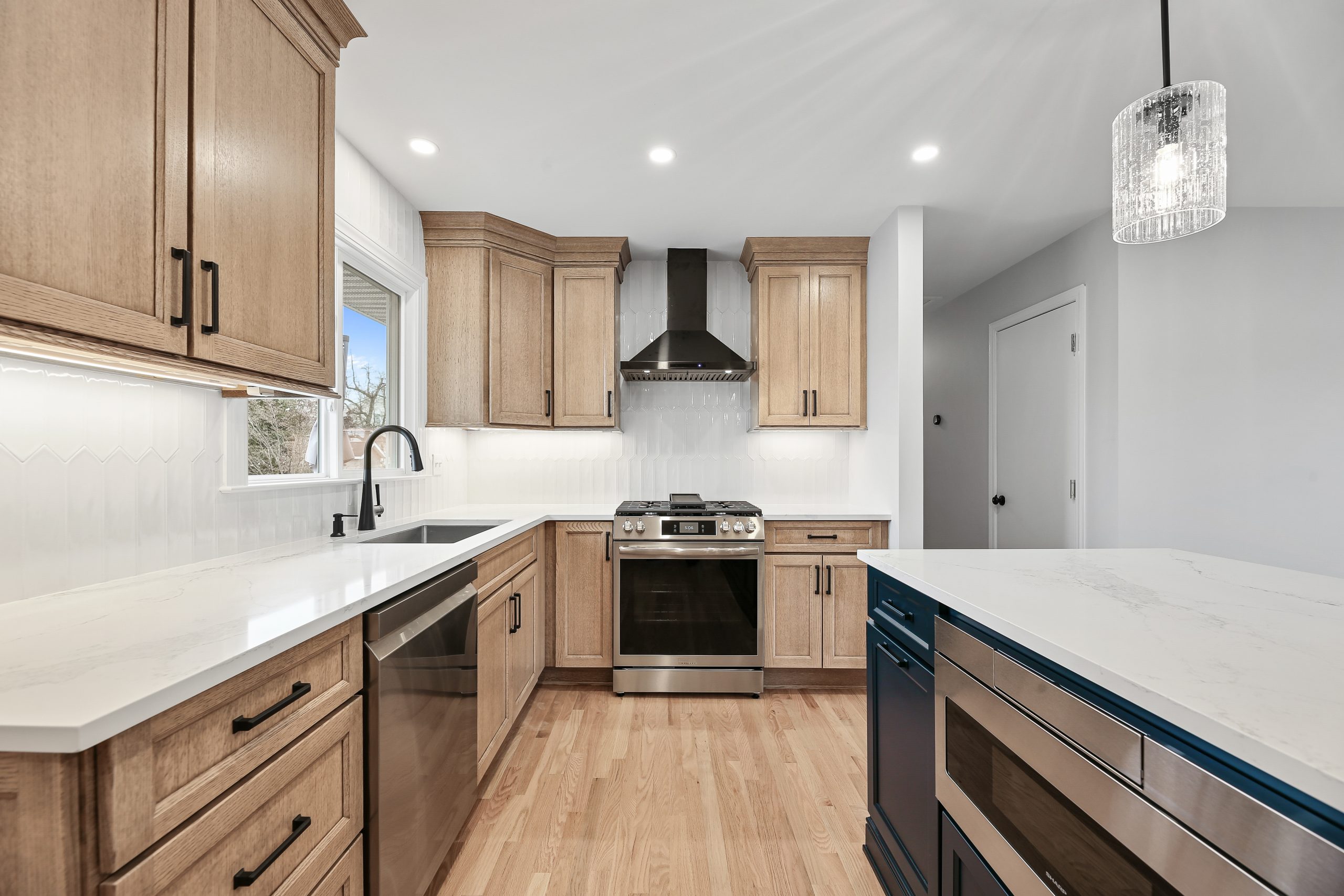 Modern kitchen with light wood cabinets, stainless steel appliances, a center island with a microwave, white marble countertops, a black range hood, and a pendant light. Hardwood floors and white tile backsplash are visible.