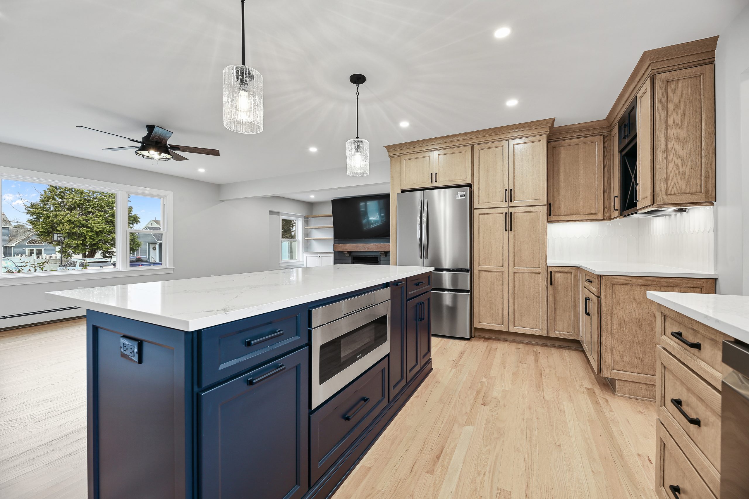 Modern kitchen with light wood cabinets, a large island with dark blue cabinetry and built-in microwave, stainless steel appliances, pendant lights, and large windows letting in natural light.