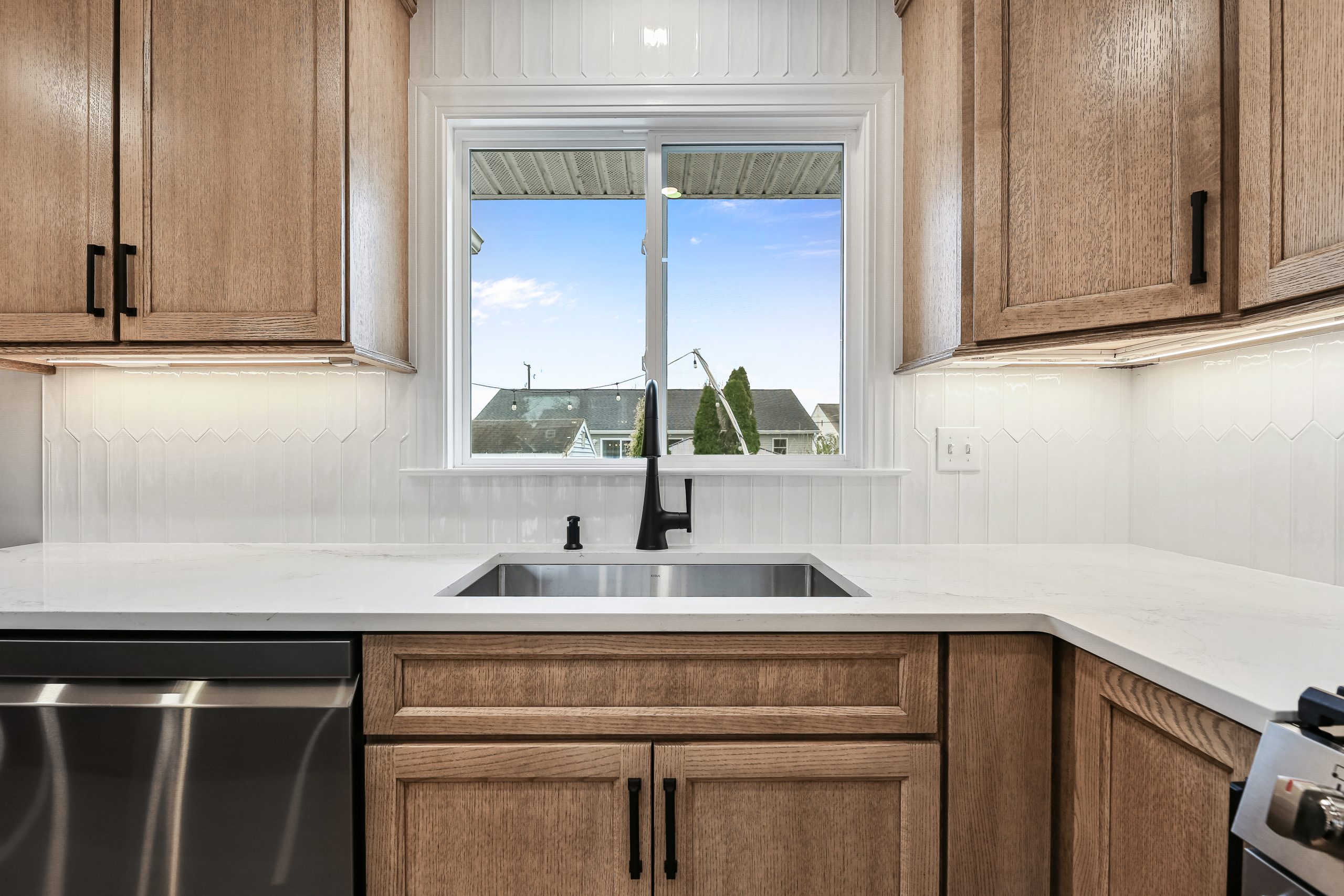 Modern kitchen with light wood cabinets, white countertops, and a stainless steel dishwasher. A sink with a black faucet is centered below a window that provides a view of houses and a clear blue sky outside.
