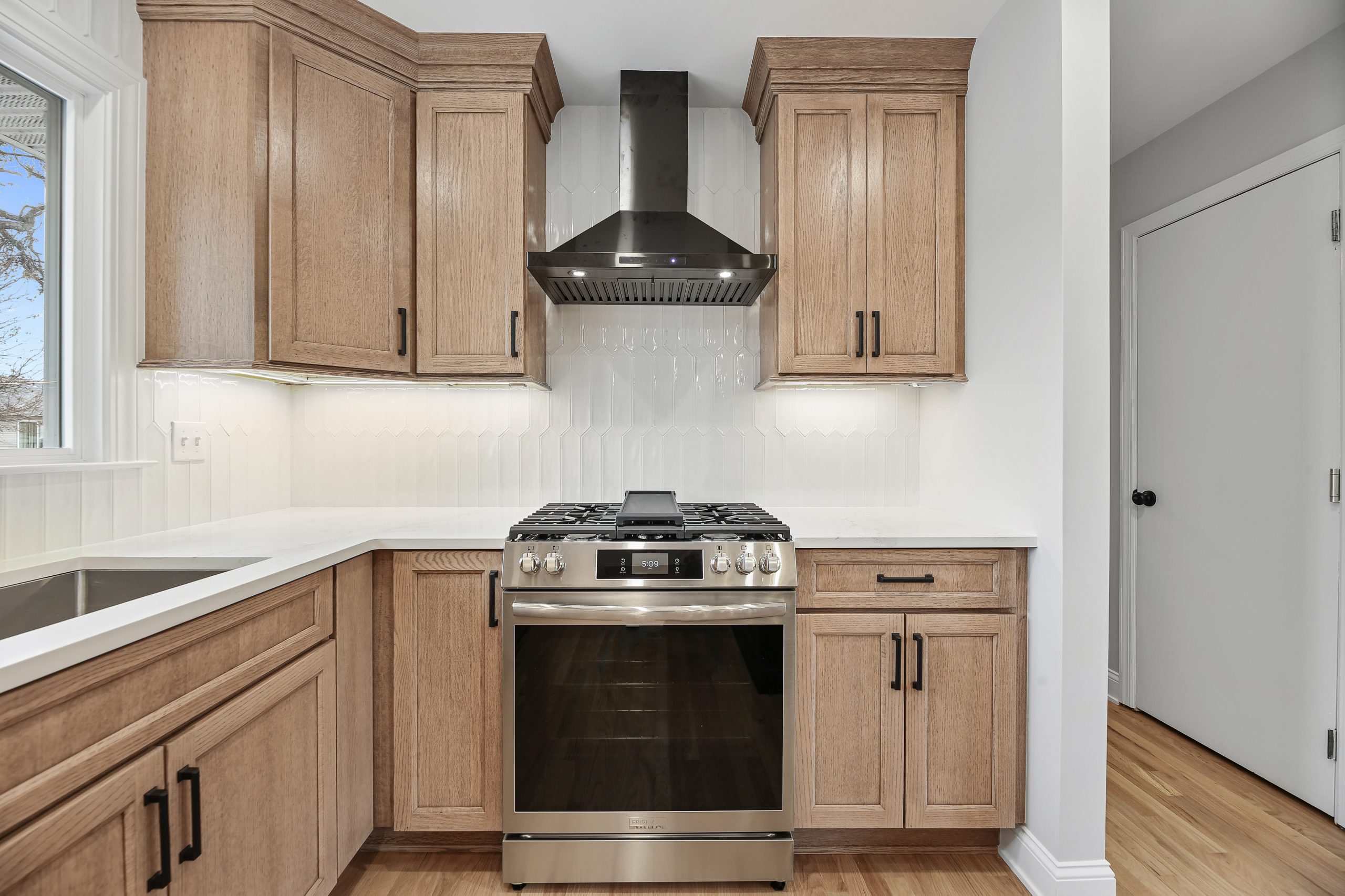 A modern kitchen with light wood cabinets, a stainless steel gas stove and oven, a black range hood, white tile backsplash, and light-colored countertops, with natural light coming from a window on the left.