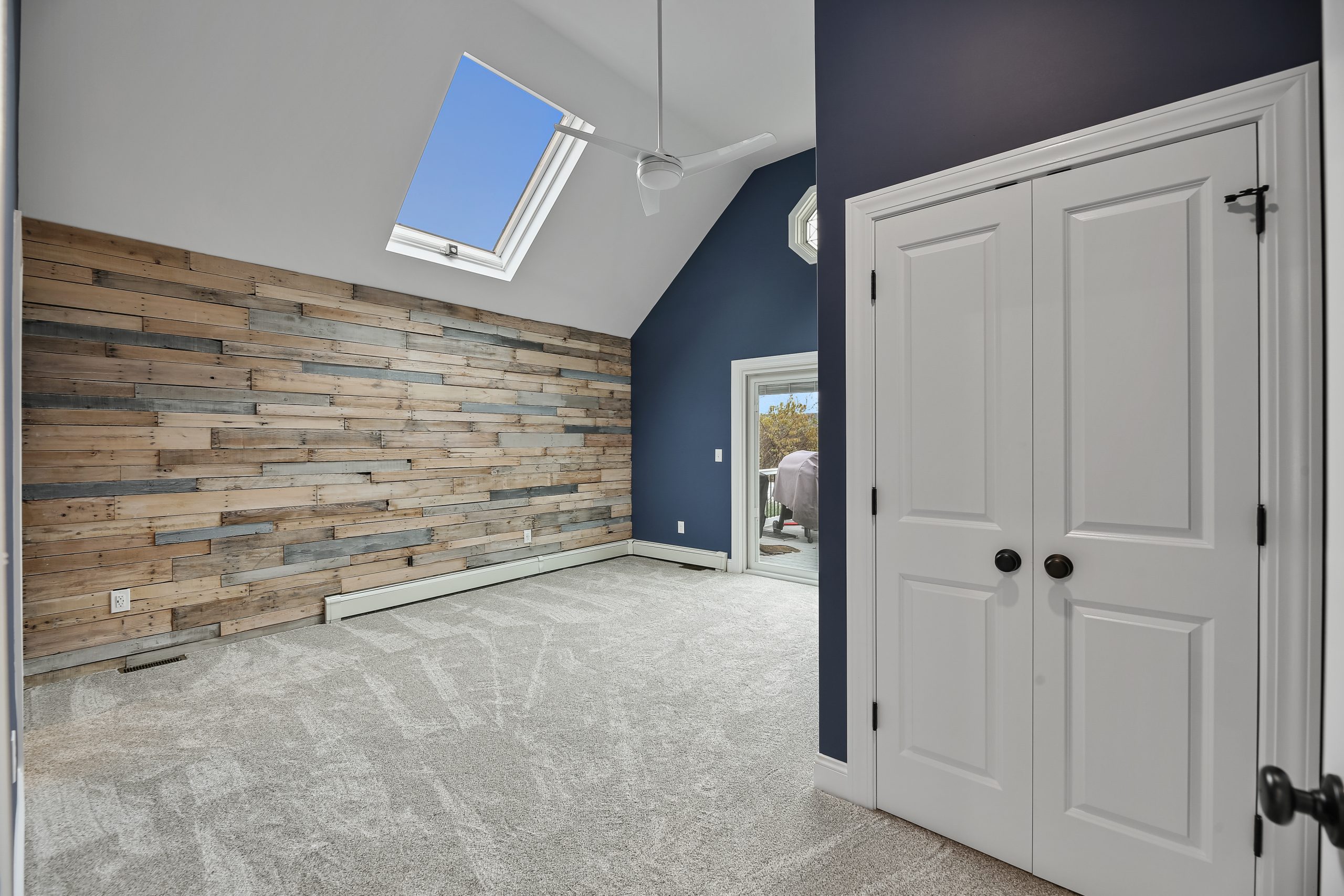 A modern empty room with a skylight, blue accent wall, double white doors, neutral carpet, and a feature wall of horizontal reclaimed wood planks. Natural light enters from the skylight and a glass door.