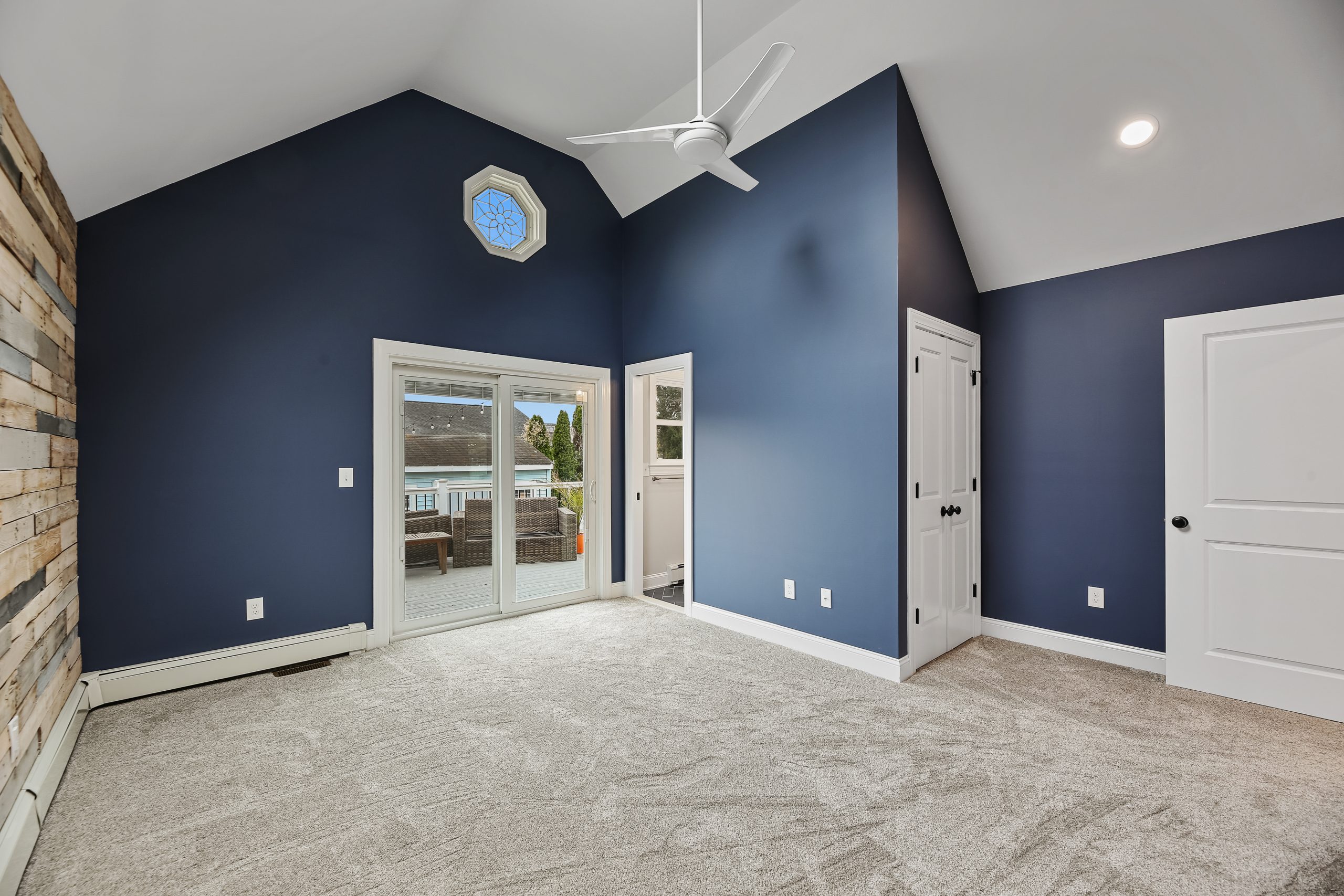 Modern, empty bedroom with navy blue walls, vaulted ceiling, ceiling fan, light gray carpet, sliding glass doors leading to a balcony, wood accent wall, and white doors and trim. Natural light enters through windows.