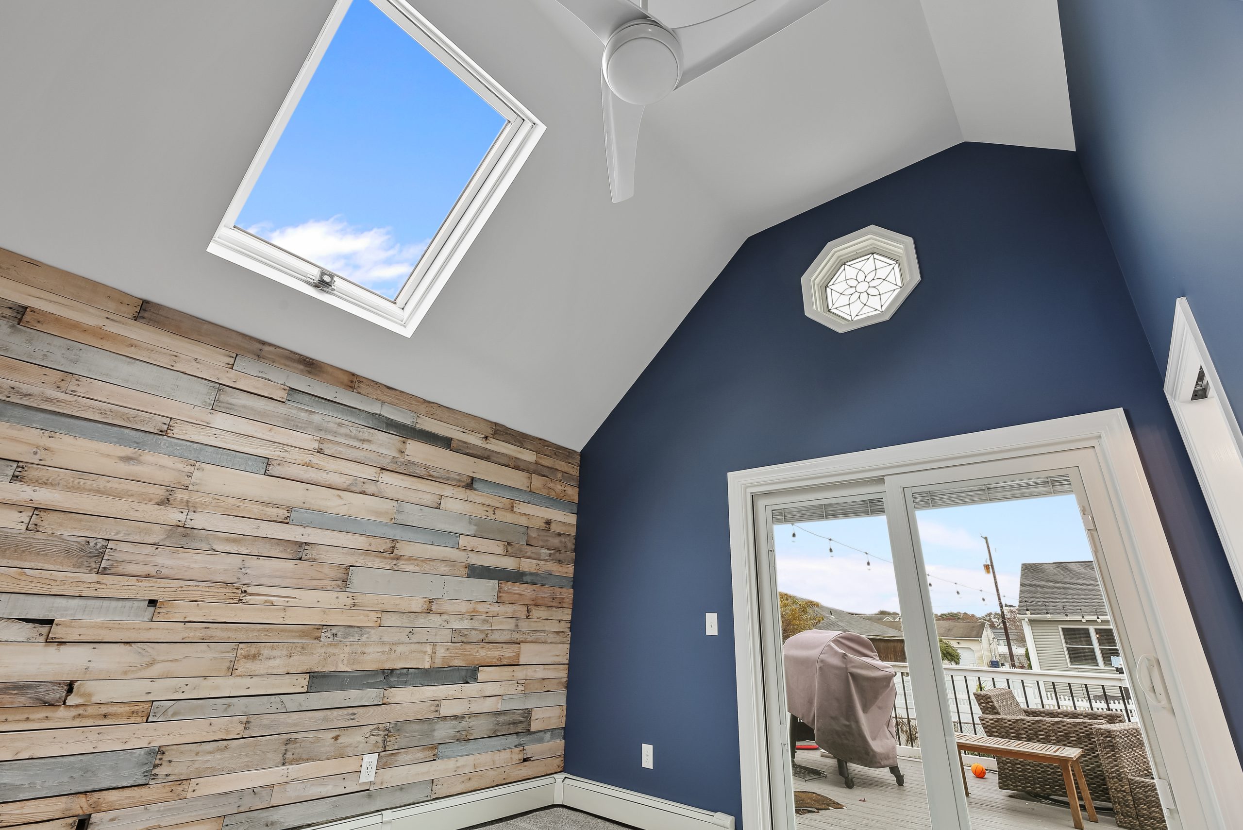 A bright room with a high vaulted ceiling, blue accent wall, reclaimed wood wall, skylight, octagonal window, and glass doors leading to a deck outside with a grill and patio furniture.