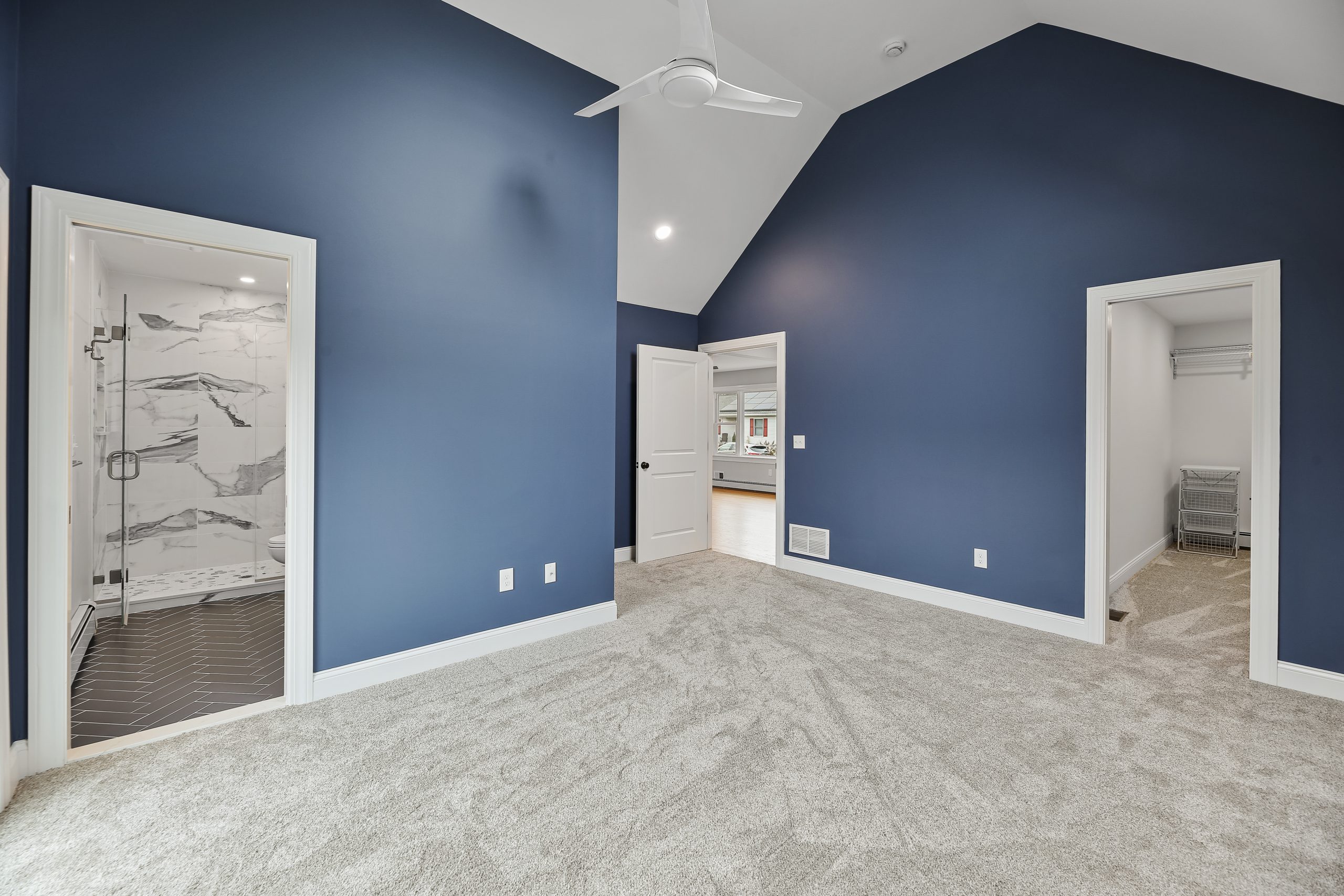 A spacious bedroom with deep blue walls, vaulted ceiling, and light carpet. The room has three doorways: one to a bathroom with marble tiles, one to a closet, and one open to a hallway.