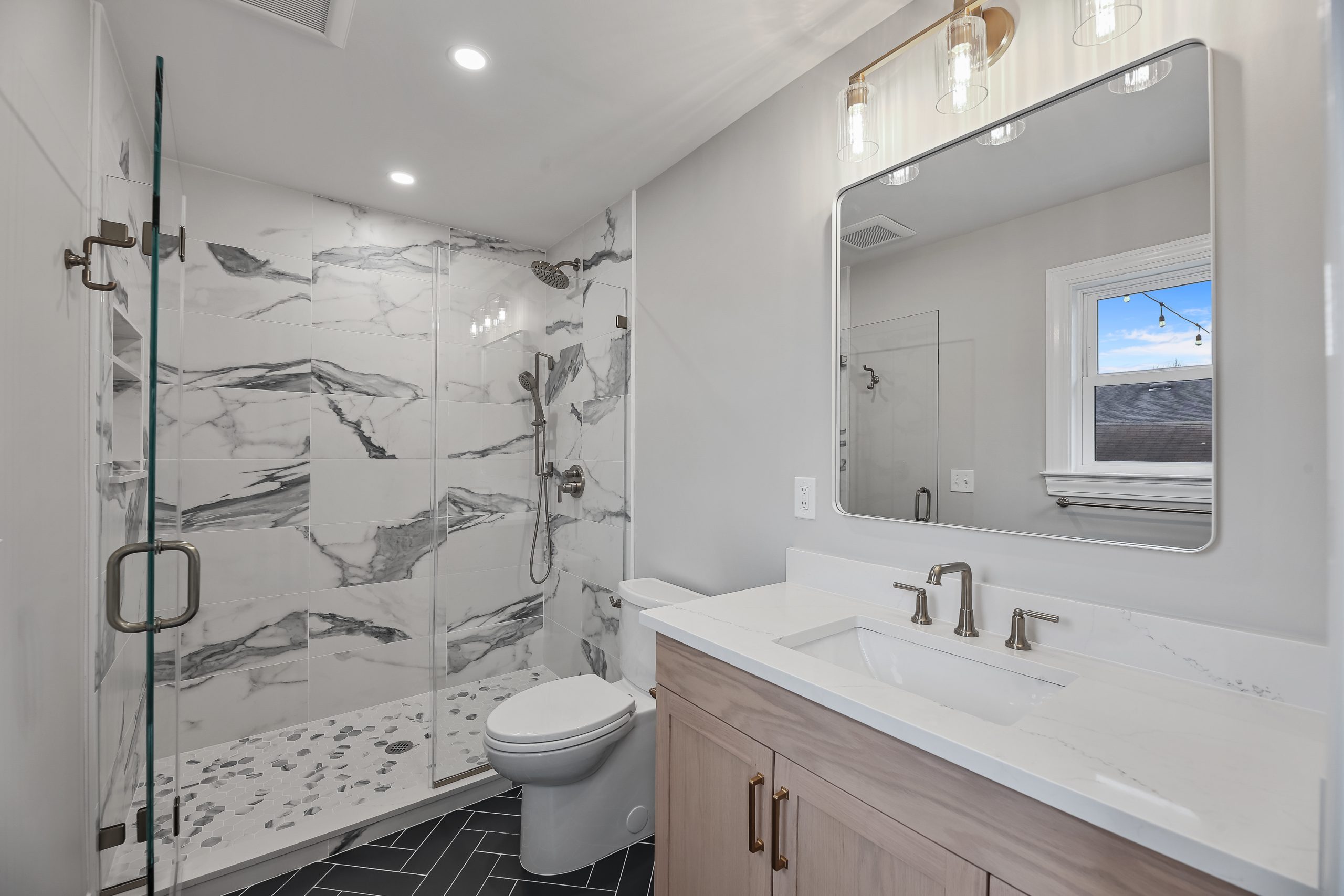 A modern bathroom featuring a glass-enclosed shower with white and gray marble walls, a toilet, a floating vanity with a white countertop, dual sinks, and a large mirror with two lights above it.