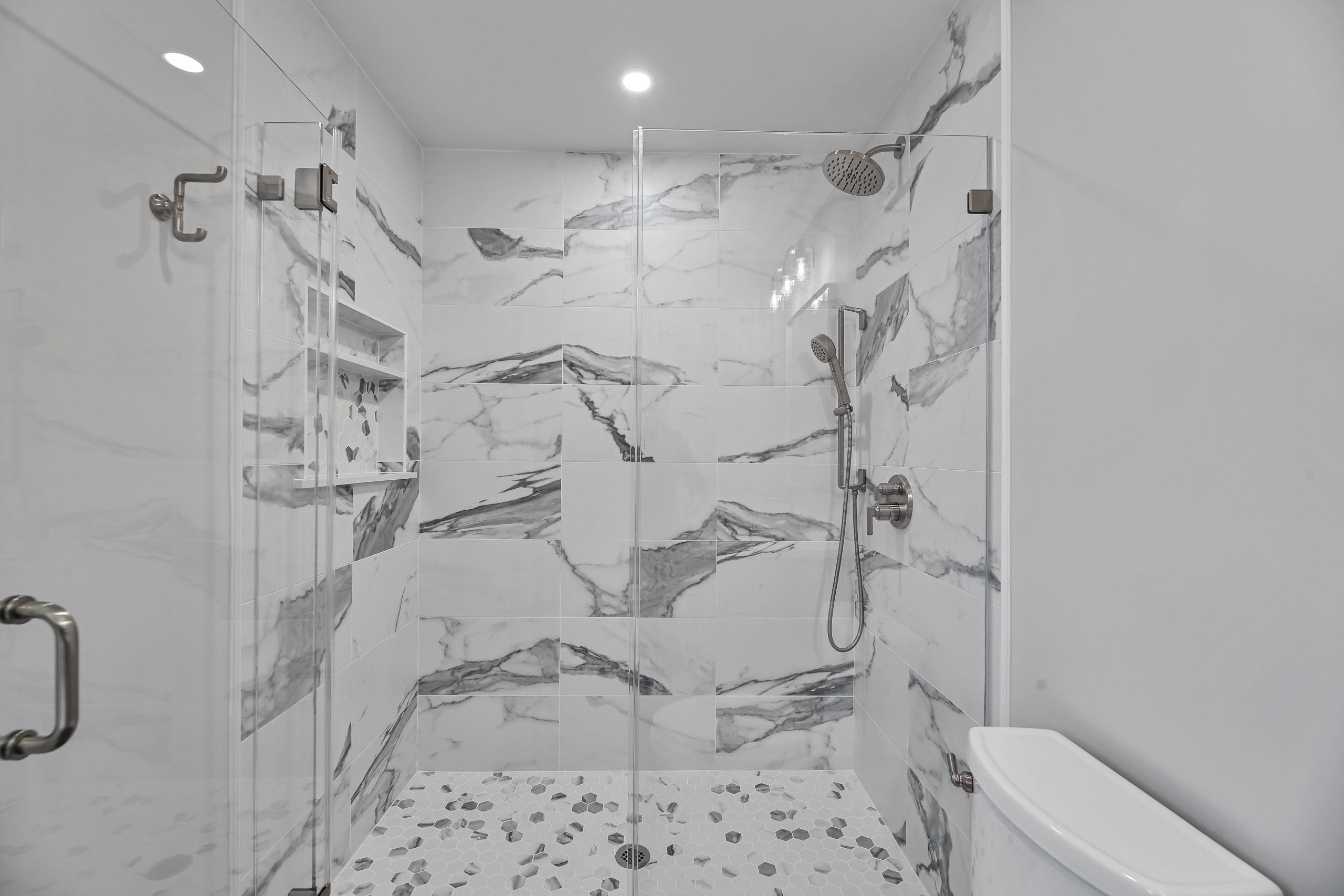 Modern walk-in shower with large white and gray marble tiles, glass doors, a rainfall showerhead, a handheld shower, built-in shelves, and a white toilet partially visible on the right.