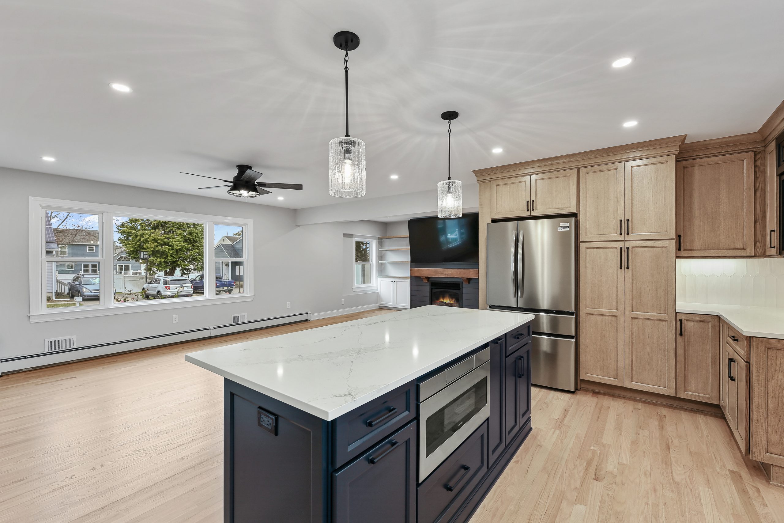 Modern kitchen with a large marble island, navy blue base, built-in microwave, stainless steel fridge, light wood cabinets, pendant lights, and open living area with fireplace and large windows.