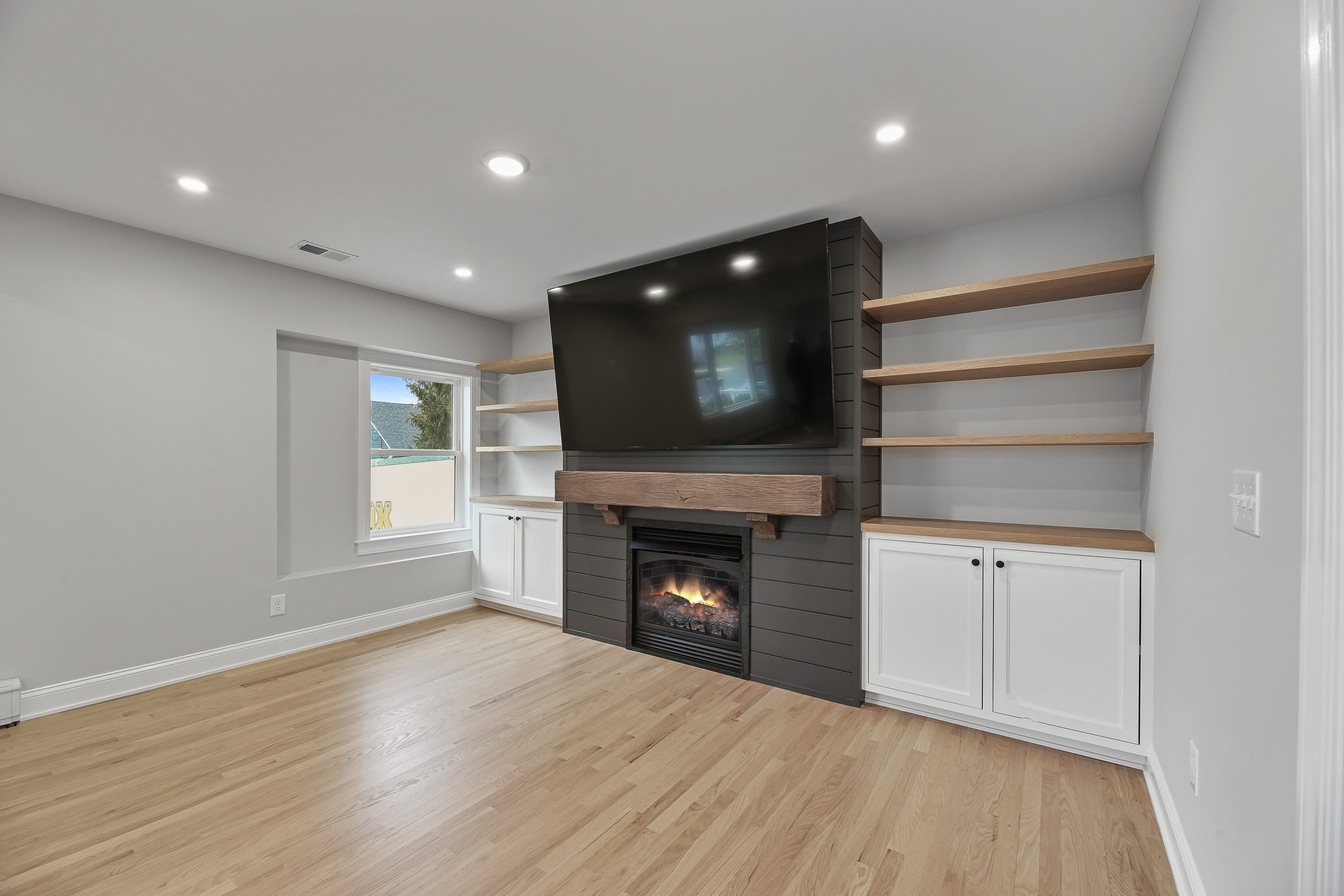 A modern living room with light wood flooring, a large TV mounted above a fireplace, built-in shelves with wooden tops on both sides, white cabinets below, and a window letting in natural light.