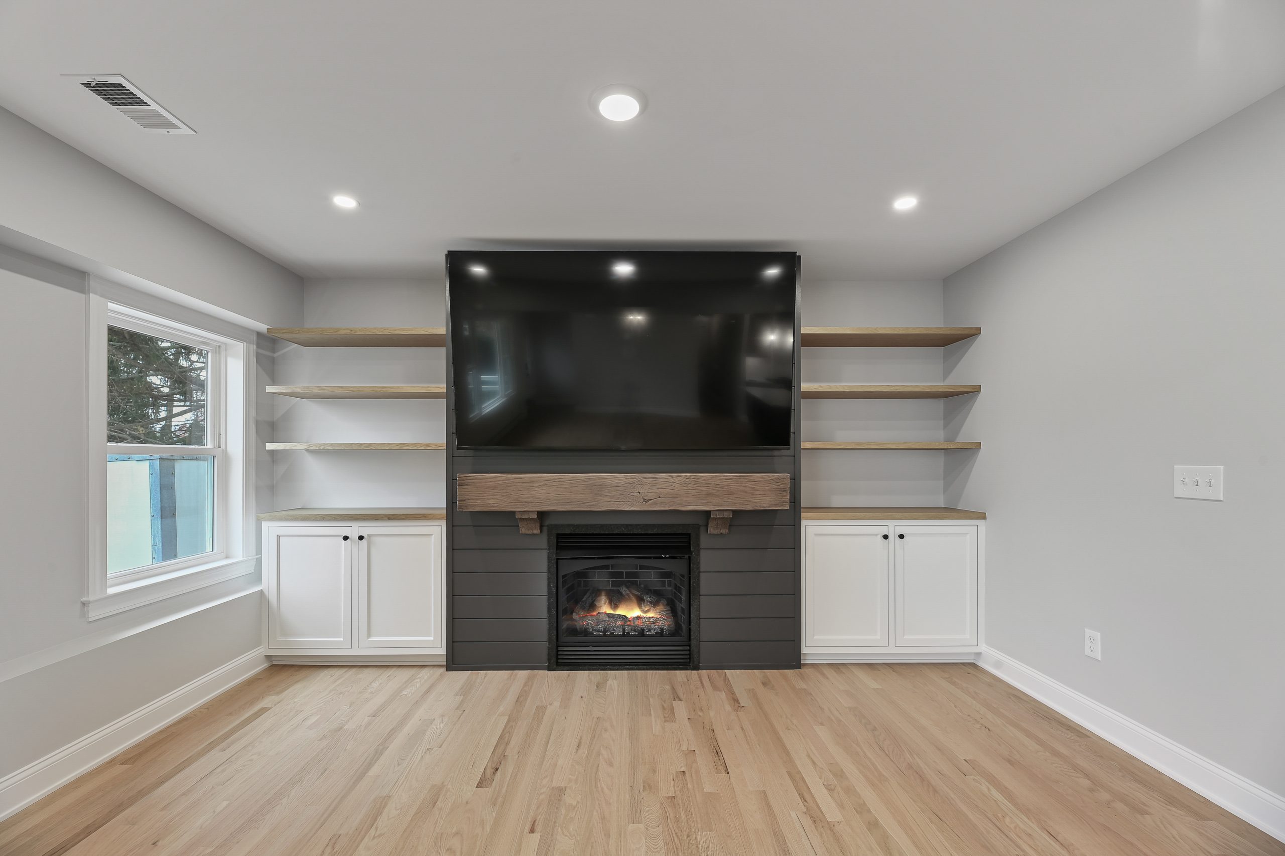 Modern living room with light wood floors, a wall-mounted TV above a fireplace, built-in shelves with white cabinets on both sides, and neutral gray walls. The room has recessed lighting and a large window on the left.