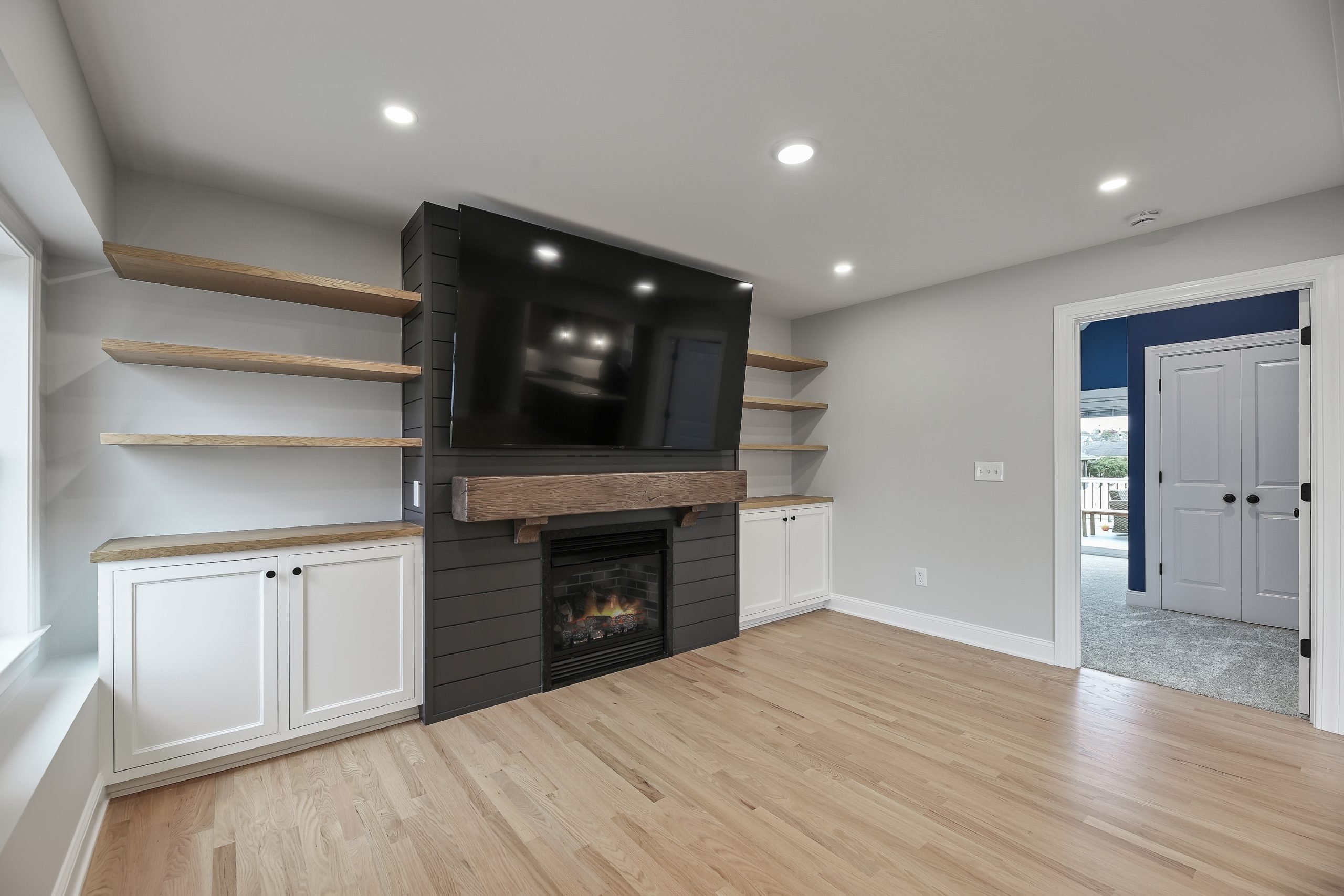 A modern living room with light wood floors, built-in white cabinets, floating wood shelves, a black fireplace, and a large flat-screen TV mounted above the mantel. White walls and recessed lighting brighten the space.
