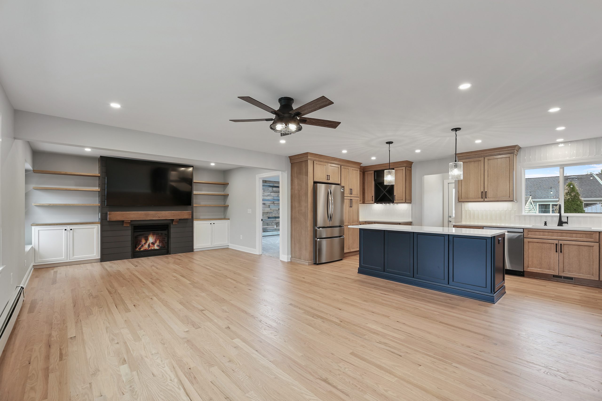 Open-concept kitchen and living space with light wood floors, modern appliances, a blue island, built-in shelves, a fireplace under a large TV, and ceiling lights with a ceiling fan.