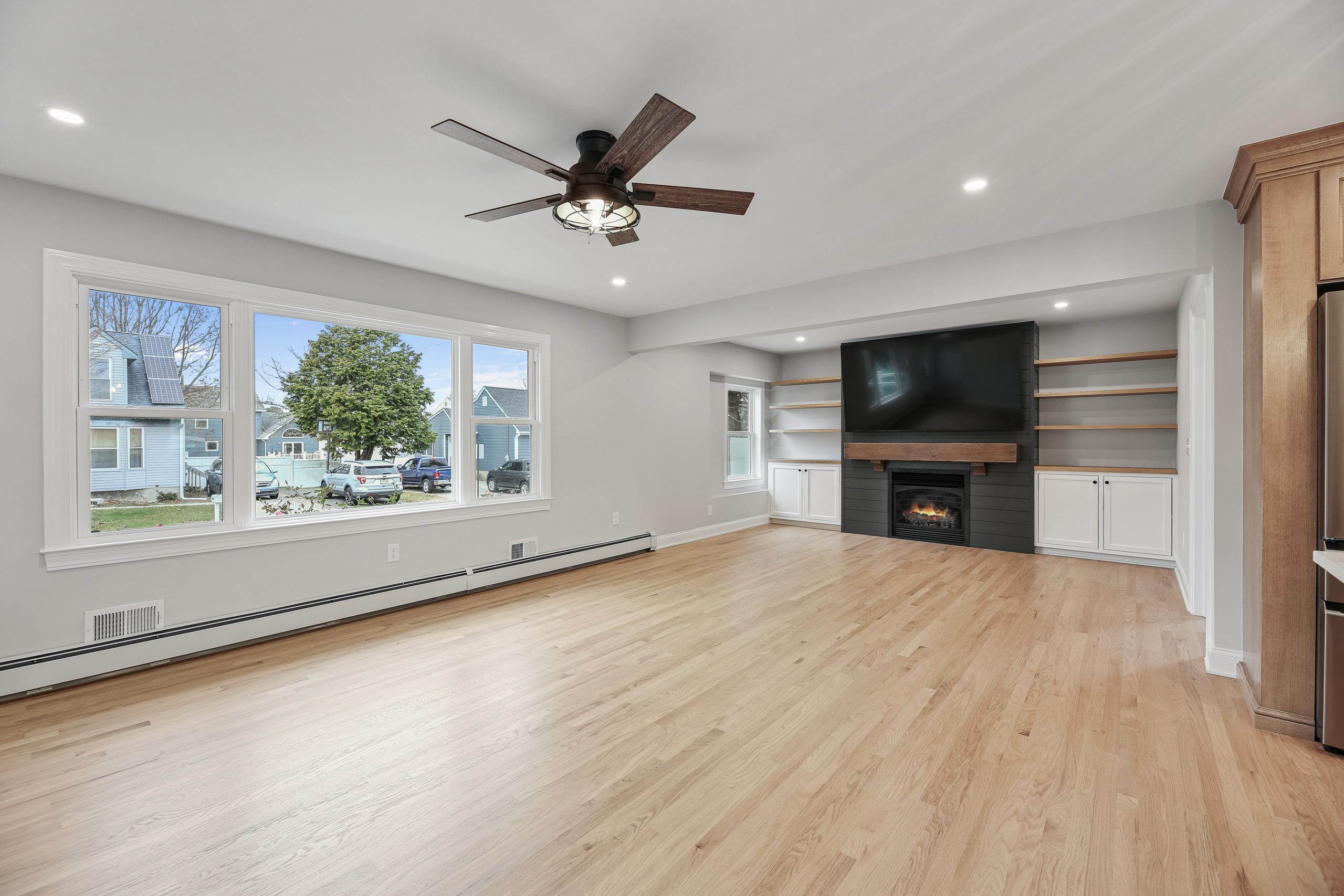 A spacious, bright living room with light wood floors, large windows, a ceiling fan, built-in shelves, a fireplace, and a mounted TV. Houses and trees are visible outside the window.