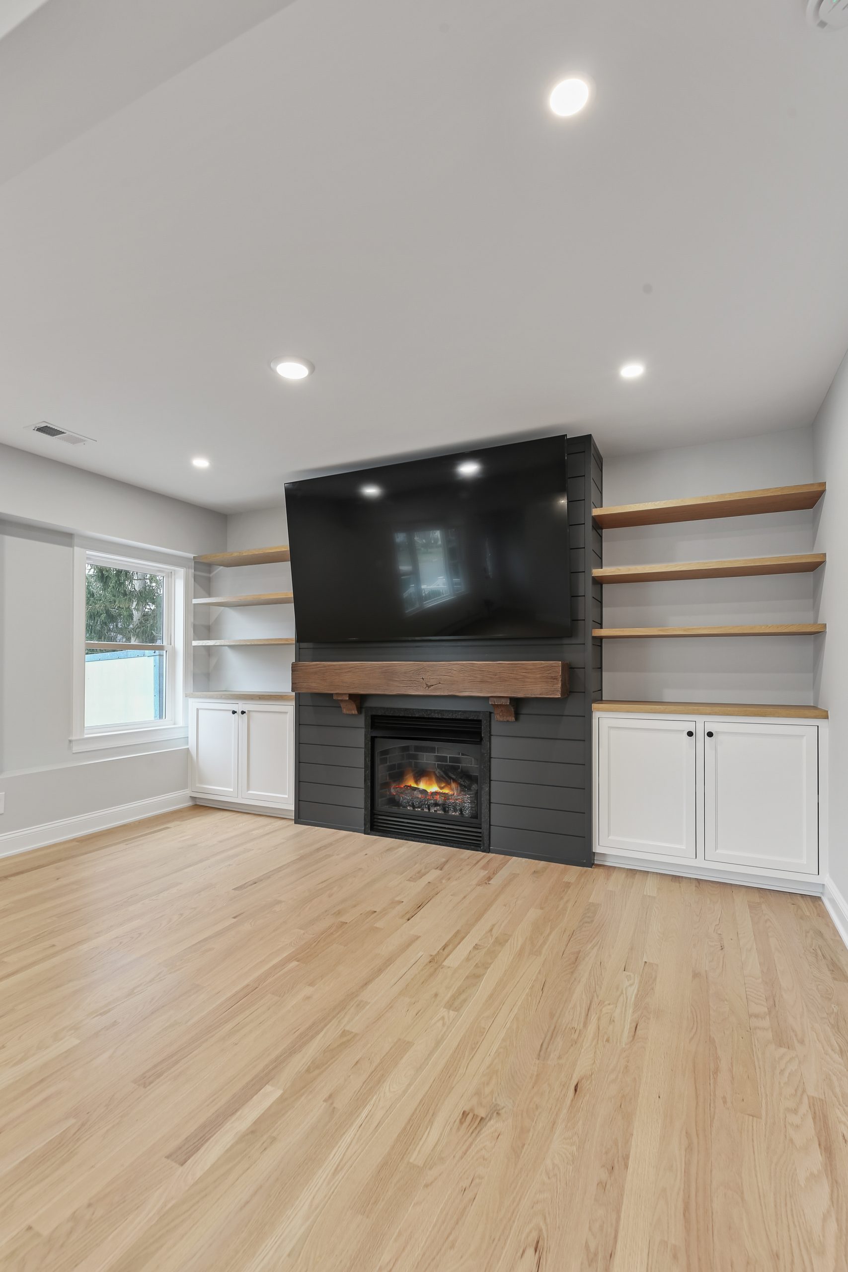 A modern living room with light wood flooring, built-in shelves, white cabinets, a wall-mounted TV above a dark fireplace, and large window letting in natural light.