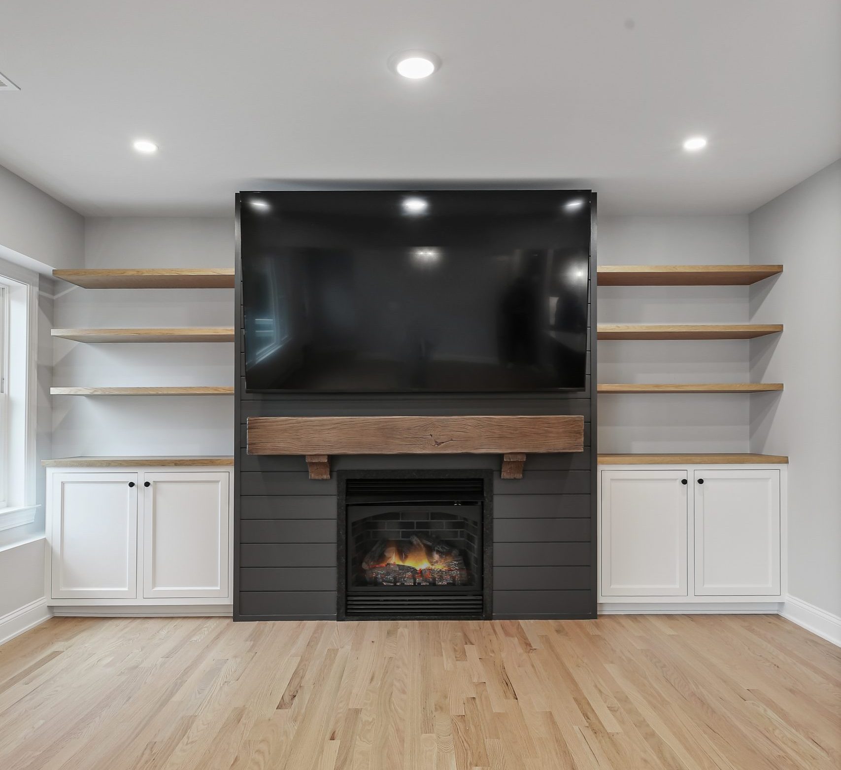 A modern living room with a large black TV mounted above a fireplace, flanked by floating wooden shelves and white cabinets on both sides, set against a light gray wall and wood flooring.