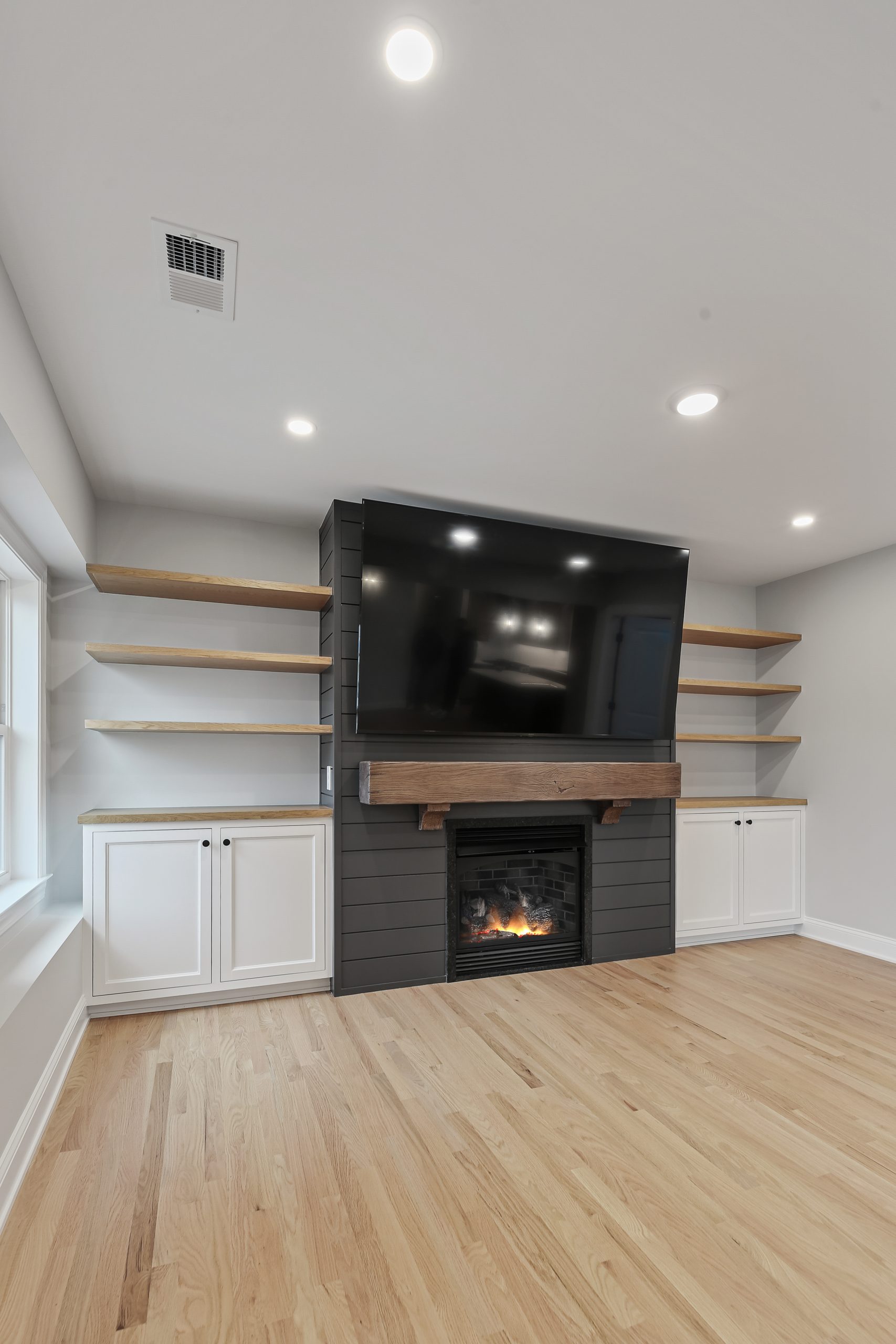 A modern living room features a wall with a large TV mounted above a fireplace, flanked by floating wood shelves and white cabinets on either side. The room has light wood floors and recessed lighting.
