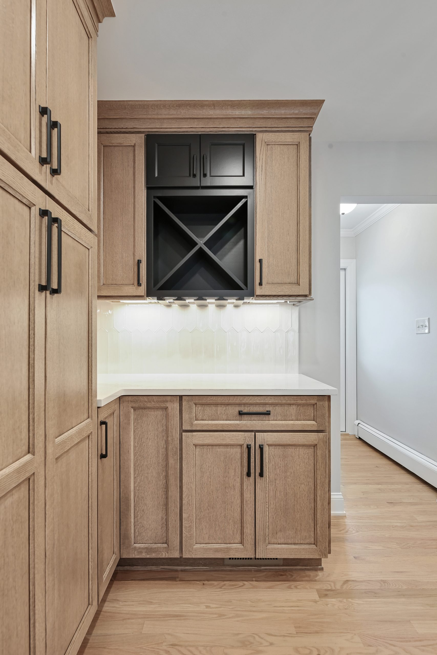 A kitchen corner with light wood cabinets, black handles, a white countertop, and a black wine rack above. The backsplash is glossy white tile, and the floor is light wood. An open doorway is visible to the right.