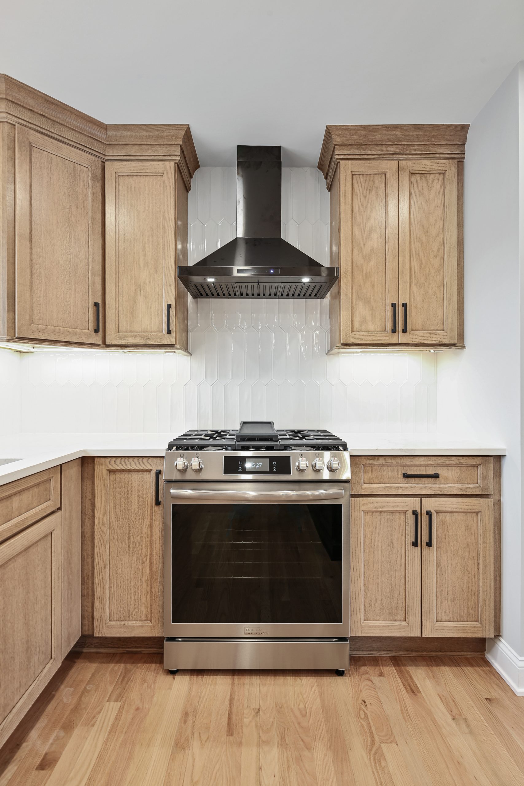 Modern kitchen with wooden cabinets, stainless steel stove and oven, black range hood, white backsplash tiles, and light wood flooring. The space is clean and well-lit with under-cabinet lighting.