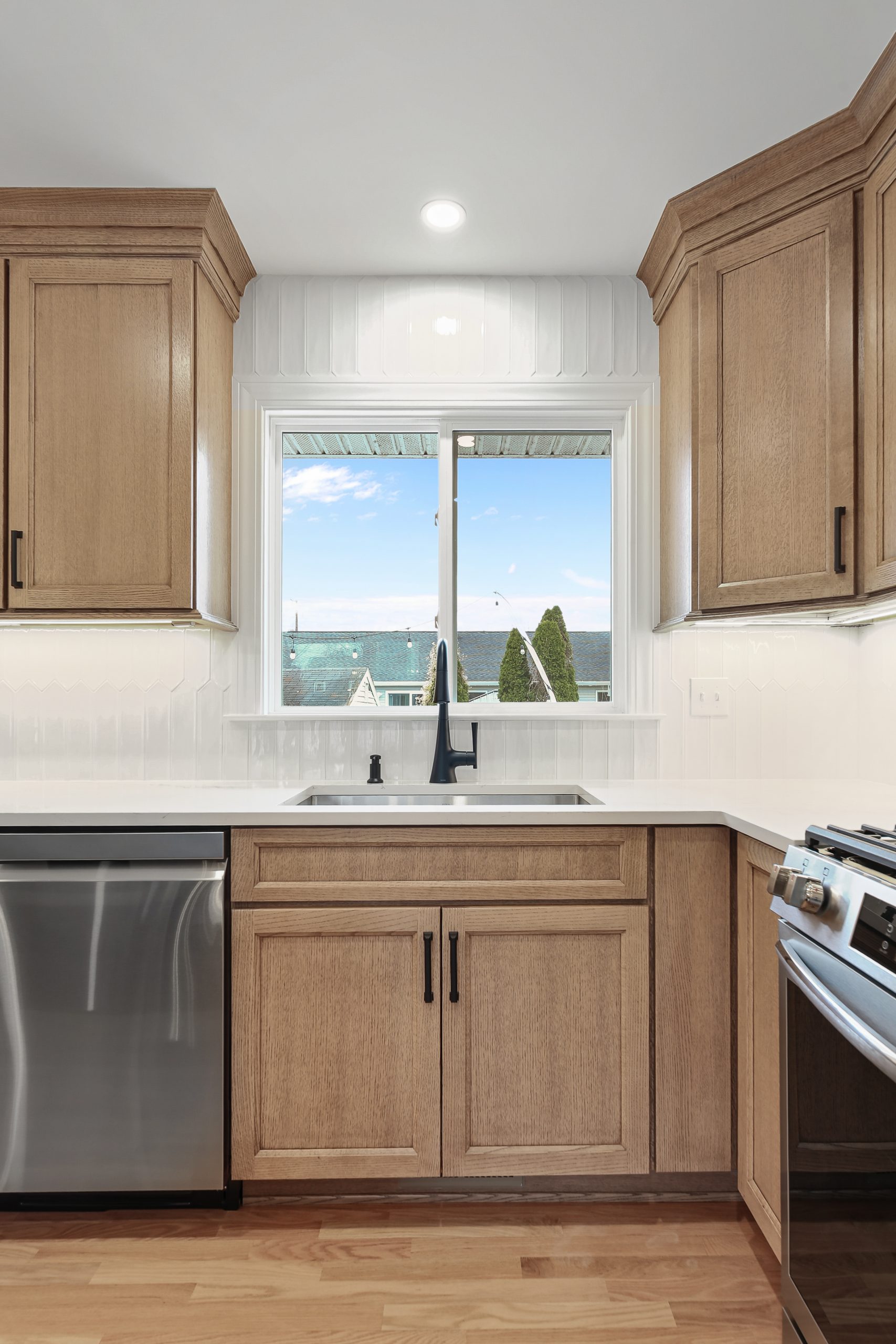 A modern kitchen with wood cabinets, white countertops, a stainless steel dishwasher, a stove, and a black faucet in front of a window showing an outdoor view with blue sky and rooftops.