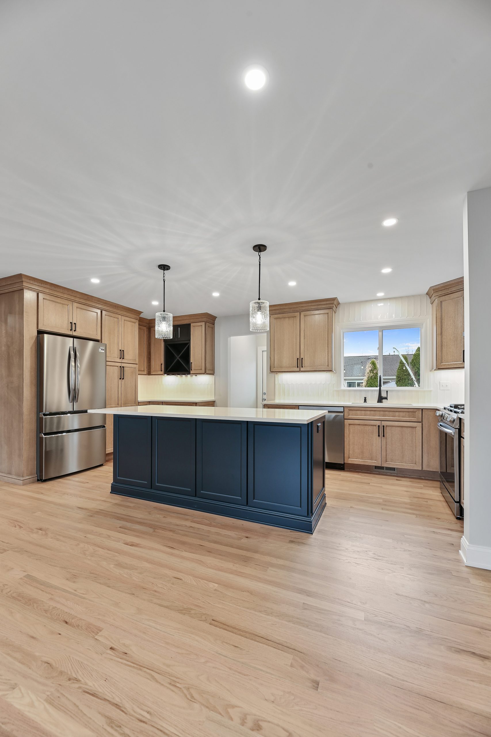 Modern kitchen with light wood cabinets, stainless steel appliances, a large navy blue island with pendant lights above, light wood flooring, and a window over the sink bringing in natural light.