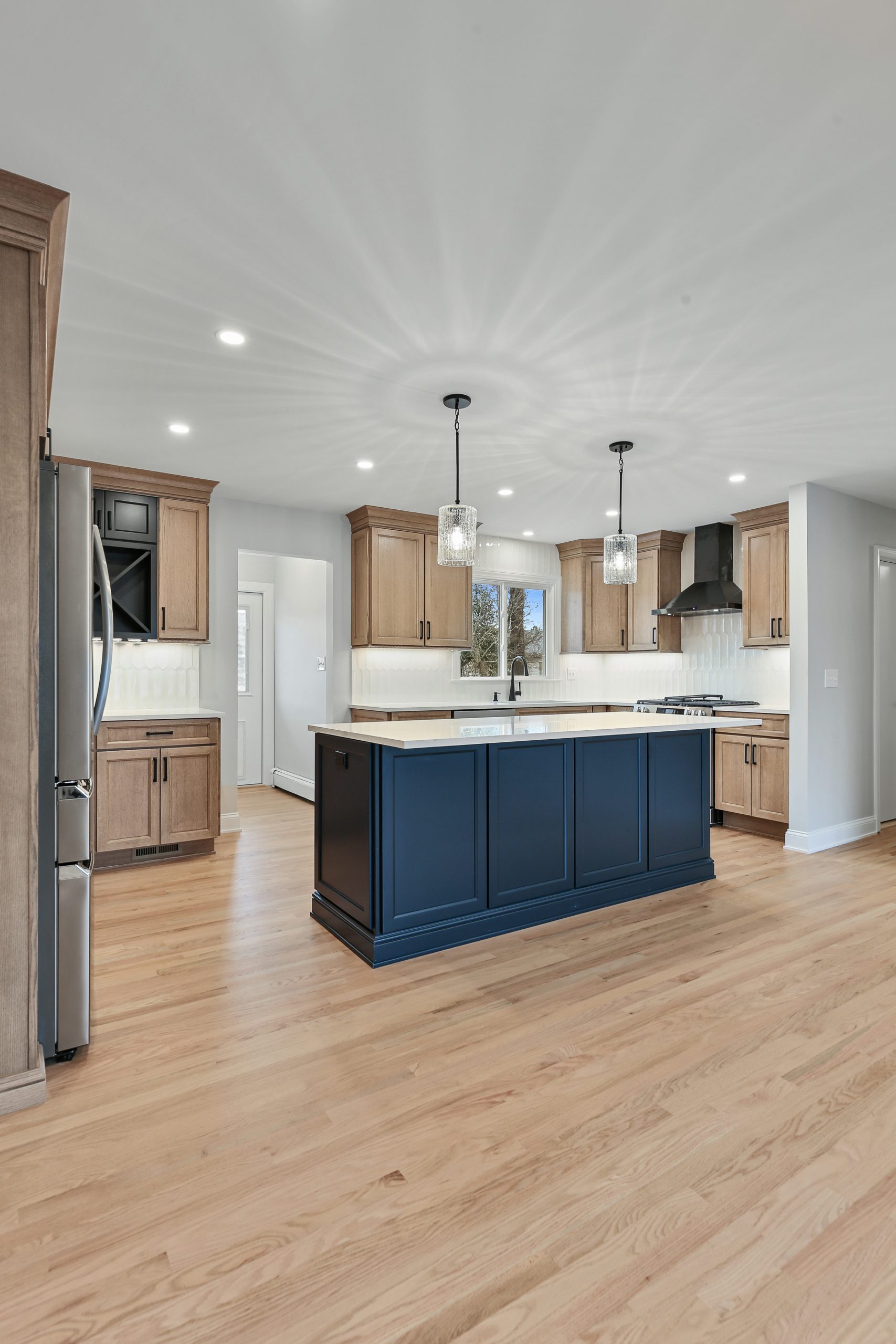 Modern kitchen with light wood cabinets, a large navy blue island, stainless steel appliances, two pendant lights, white countertops, and light hardwood floors. The space is bright with natural and recessed lighting.