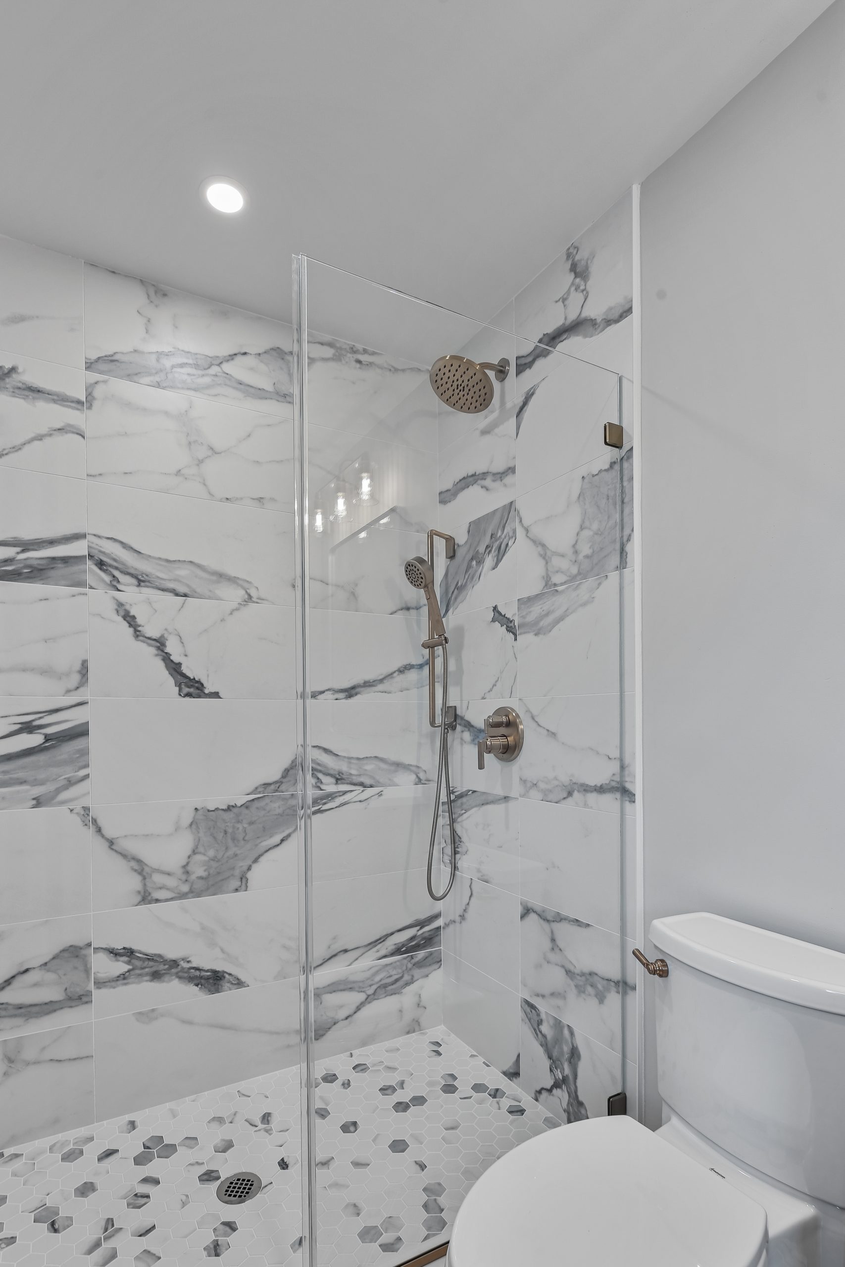 Modern bathroom with a glass-enclosed shower featuring large white and gray marble tiles, a rainfall showerhead, and a handheld sprayer. White toilet and hexagonal floor tiles are visible in the foreground.
