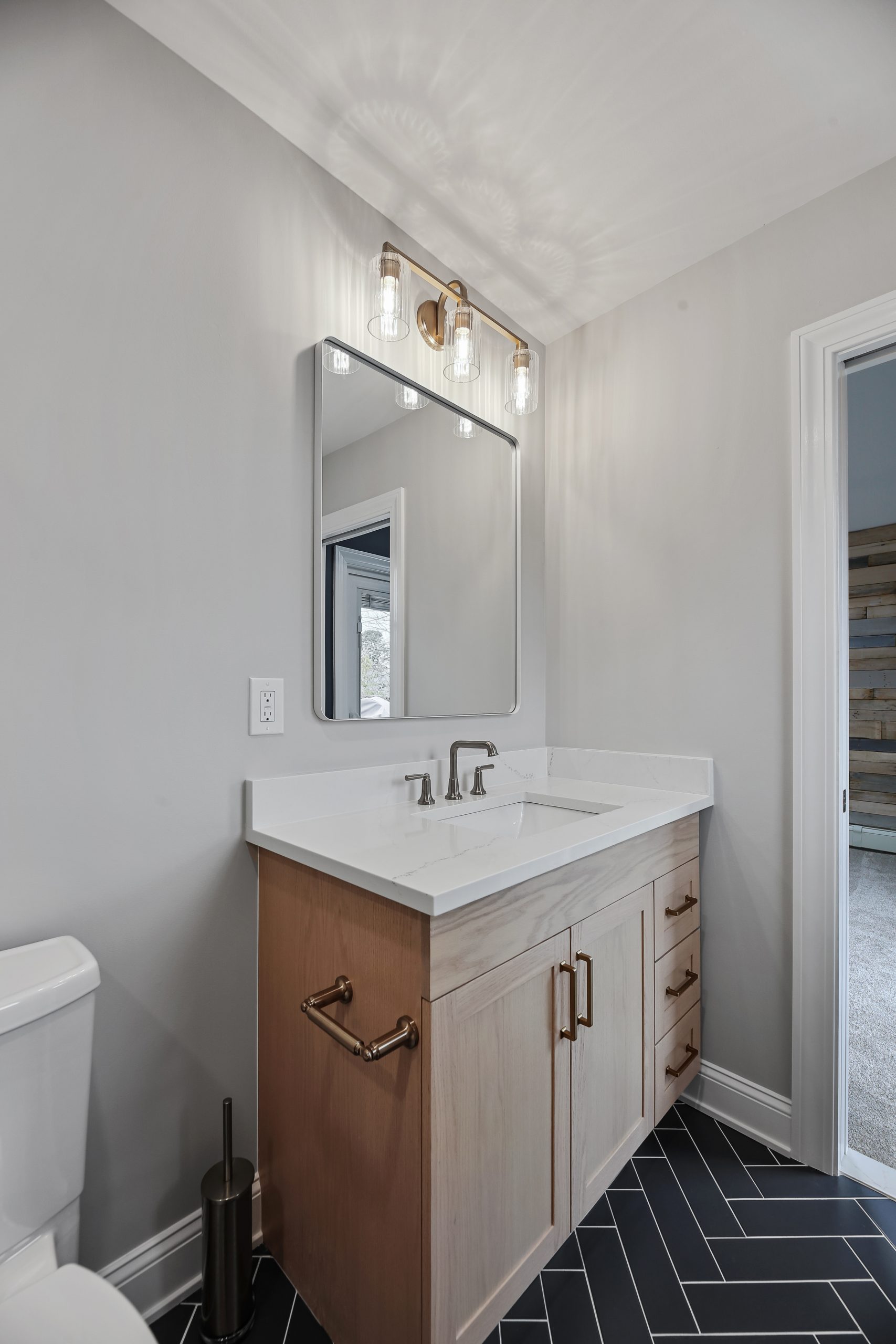 Modern bathroom vanity with a rectangular mirror, dual sink faucets, light wood cabinetry, and brass hardware. Overhead, three glass-shaded lights are mounted above the mirror. Black chevron tile covers the floor.