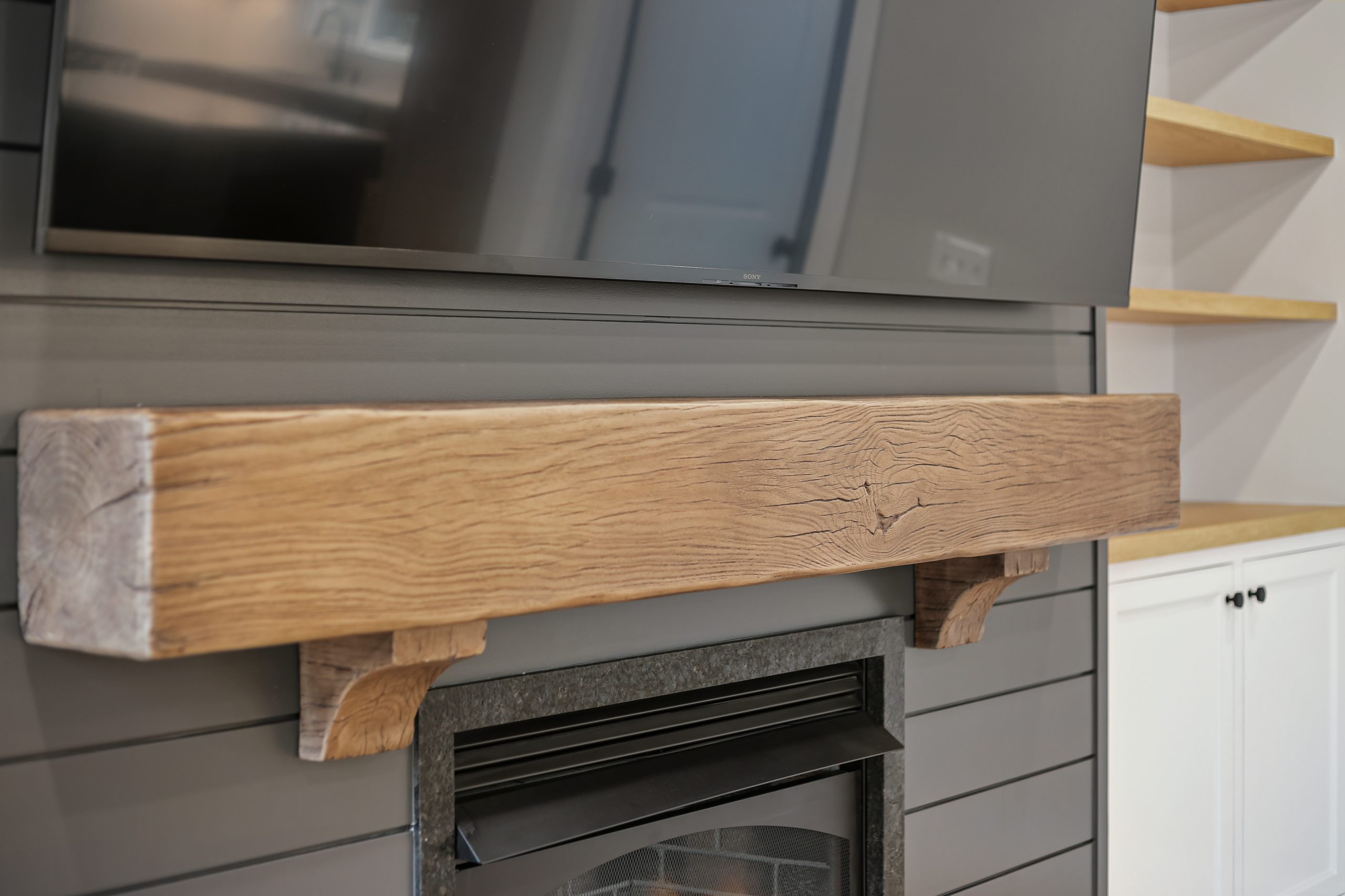 A close-up of a rustic wooden mantel above a modern fireplace, with a large flat-screen TV mounted above. The background features gray shiplap walls and floating wooden shelves on the right side.