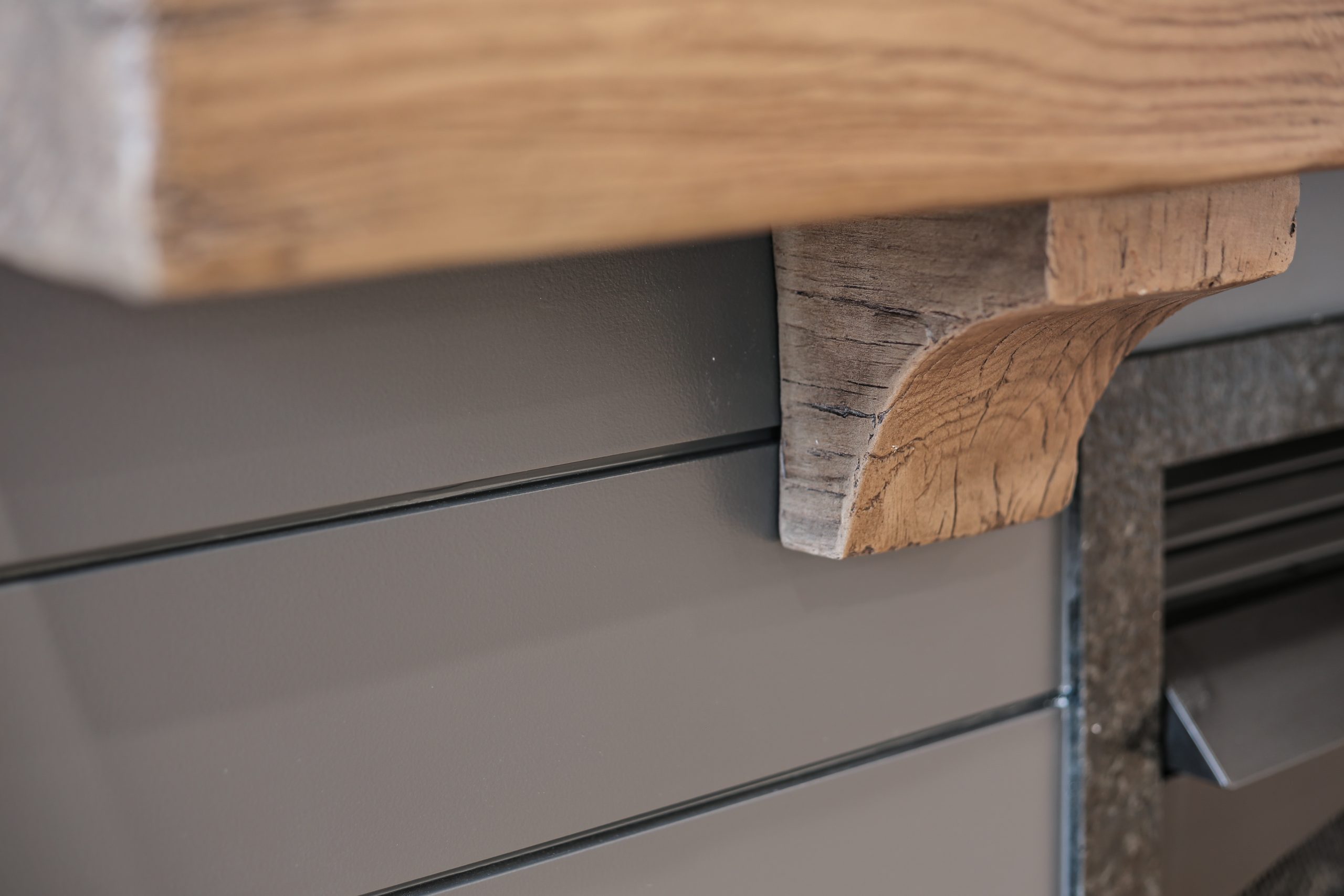 Close-up of a wooden mantel with a decorative support bracket mounted on a gray, paneled wall. The wood shows natural grain and texture, contrasting with the smooth, modern wall surface.