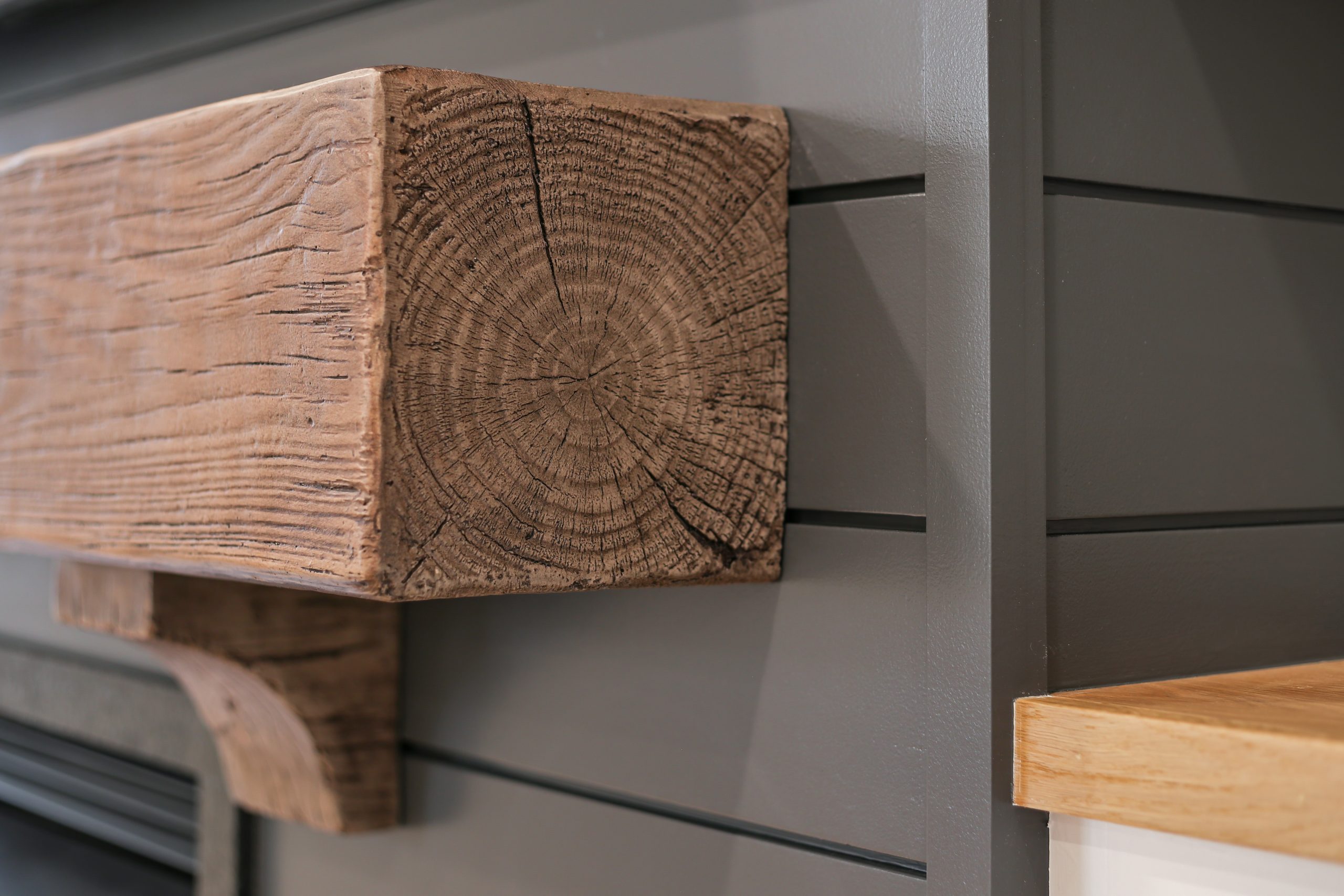 Close-up of a thick, rustic wooden mantel mounted on a gray shiplap wall, showing the detailed wood grain and texture. A lighter wood surface is partially visible in the lower right corner.