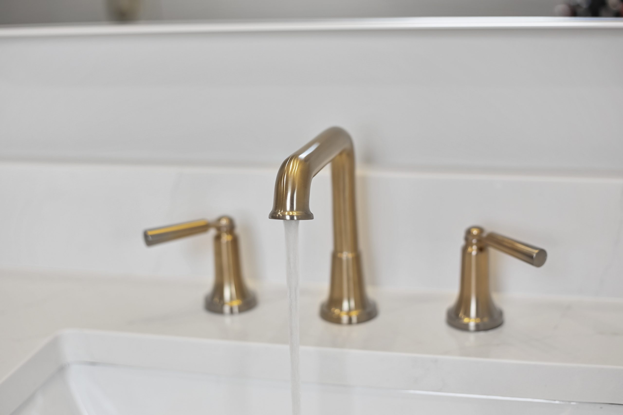 A close-up of a modern bathroom sink with a gold faucet and two matching handles, with water running from the faucet into a white basin.