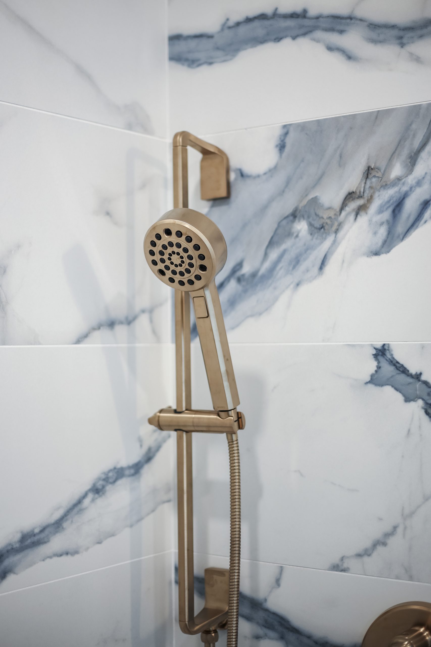Close-up of a modern gold-colored handheld showerhead mounted on a vertical bar, set against white tiles with blue-gray marble-like veining.