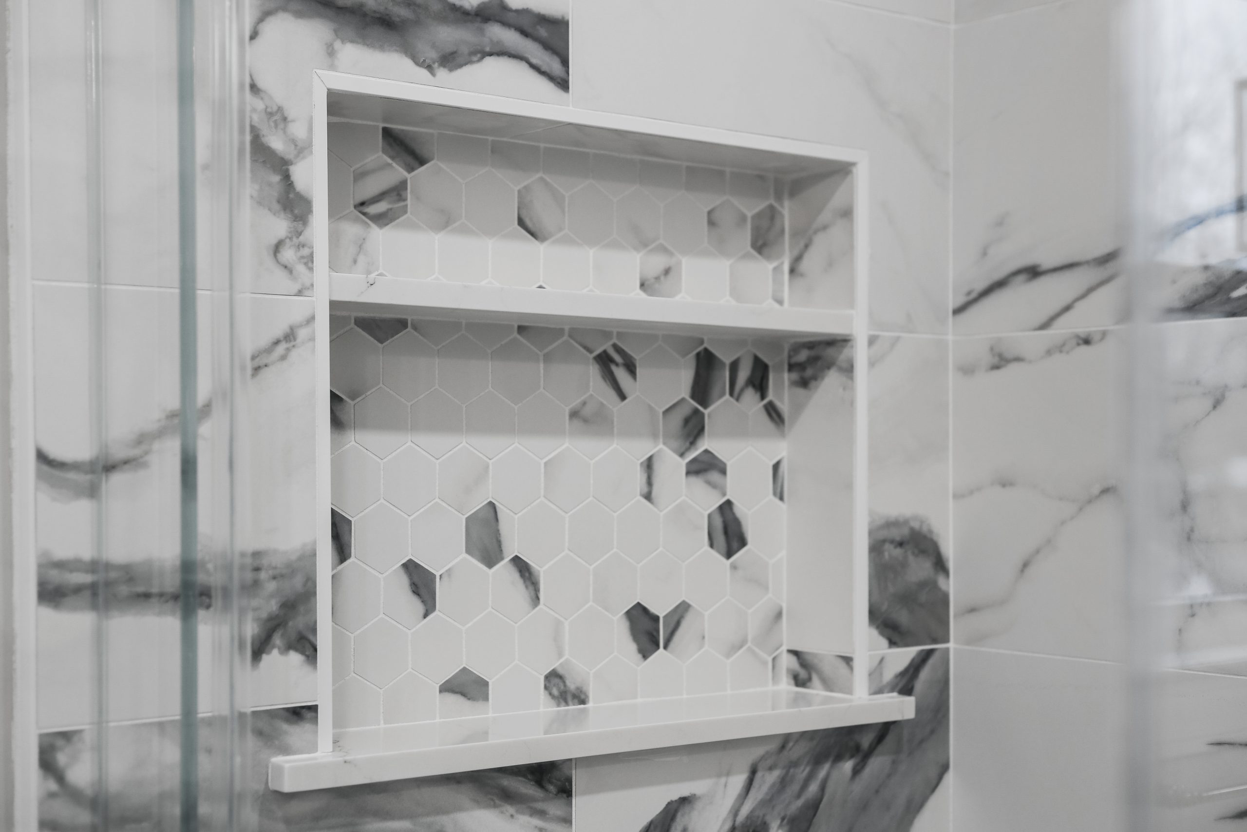 A recessed shower shelf with white hexagon tiles and scattered darker hexagon accents, set in a wall with large white and gray marble-patterned tiles.