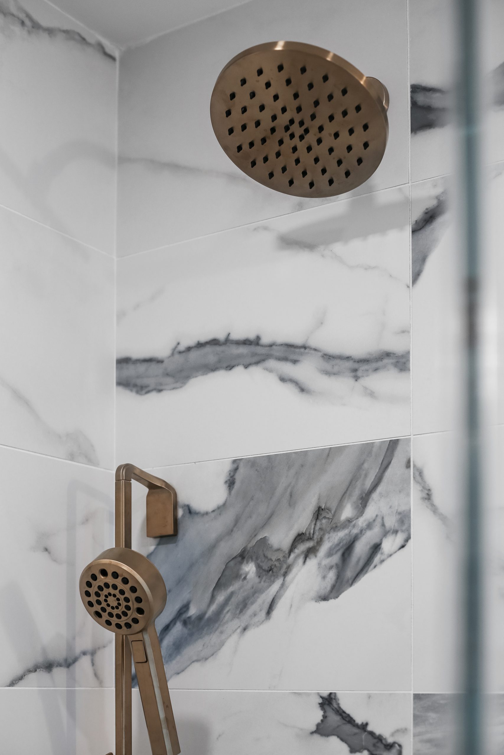 Modern shower with gold fixtures, including a large overhead rain showerhead and a handheld shower, set against white marble tiles with dark gray veining.