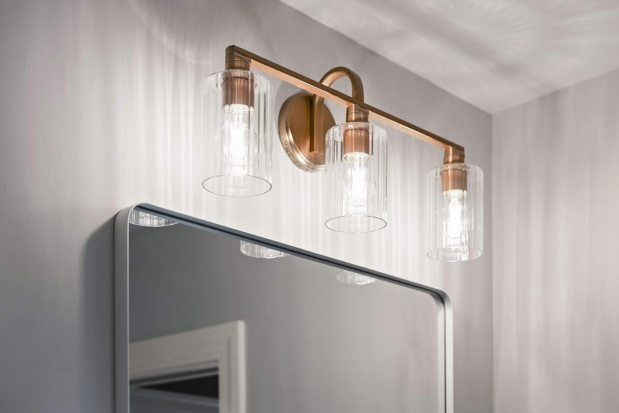 A modern bathroom light fixture with three cylindrical glass shades and a brass base is mounted above a rectangular mirror on a light gray wall. The lights are turned on, casting patterns on the wall.